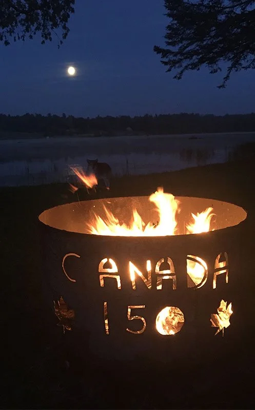 A fire pit burning with flames, decorated with cutouts spelling 'CANADA 150' and maple leaf shapes, set near a lake at night with the moon visible in the sky and a silhouette of a dog in the background.