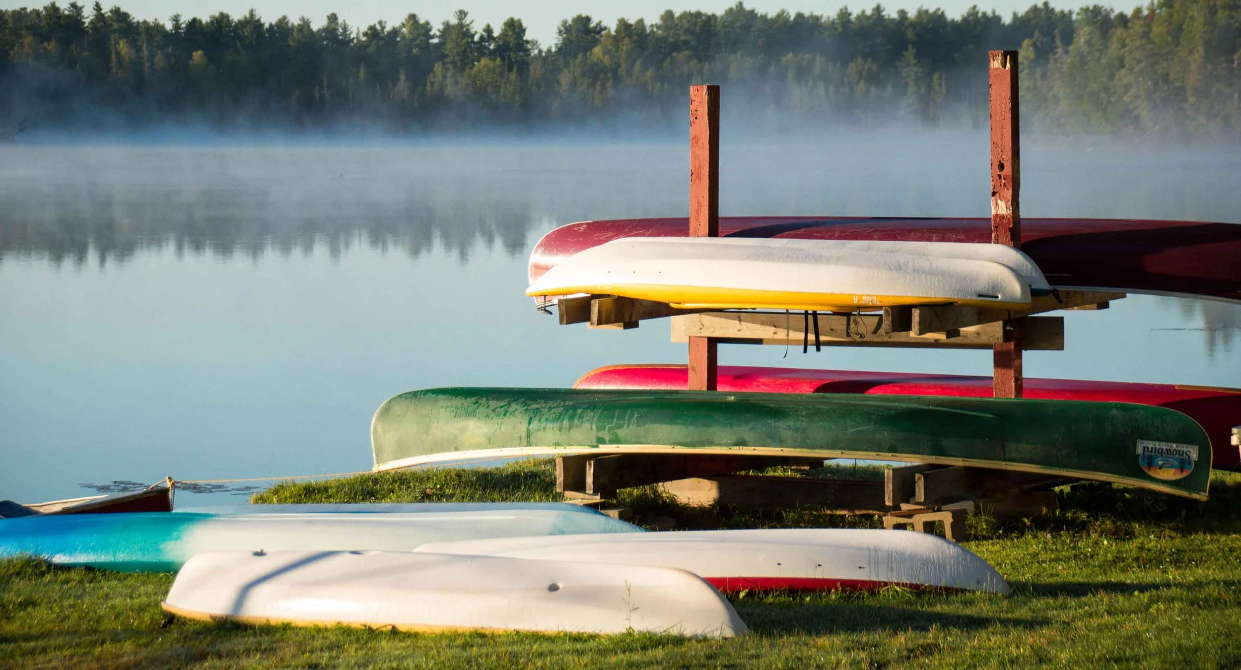Colorful kayaks and canoes stored on a wooden rack near a calm lake with mist and trees in the background.
