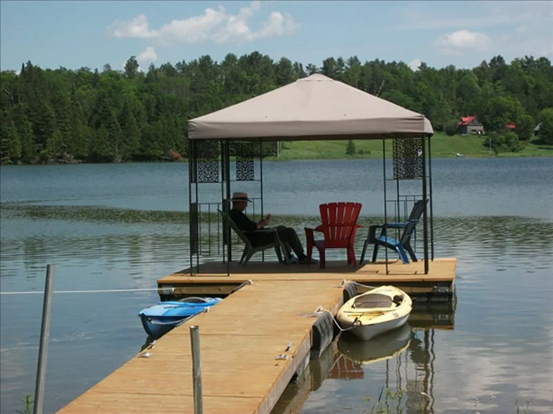 A person with a hat sitting on a chair under a gazebo on a lake dock, surrounded by boats and calm water, with trees and houses in the background.