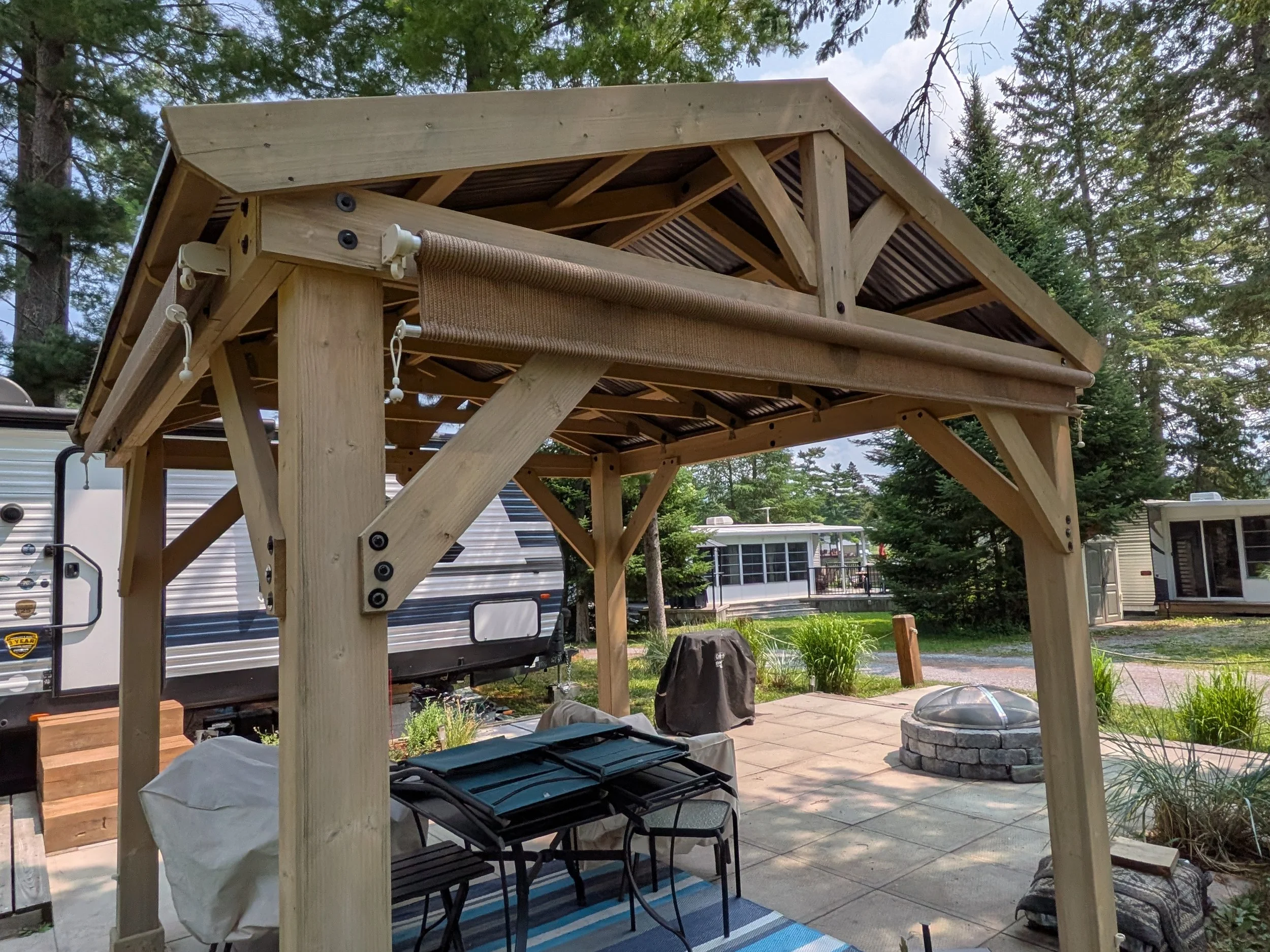 Backyard scene with a wooden deck under construction, chairs, poured concrete fire pit, RV, trees, and outdoor furniture.