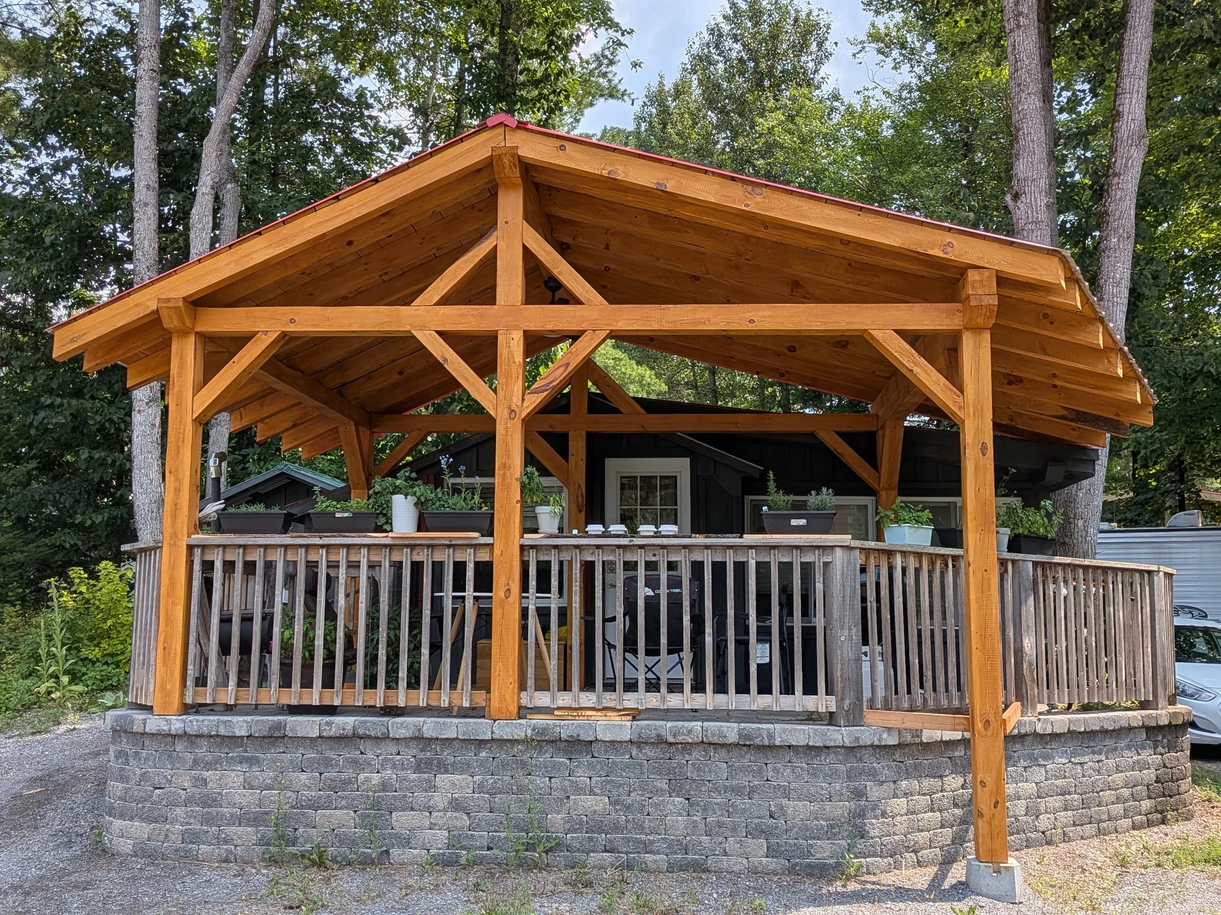 A wooden deck with railing and potted plants, covered by a wooden roof structure, situated in a backyard with trees.