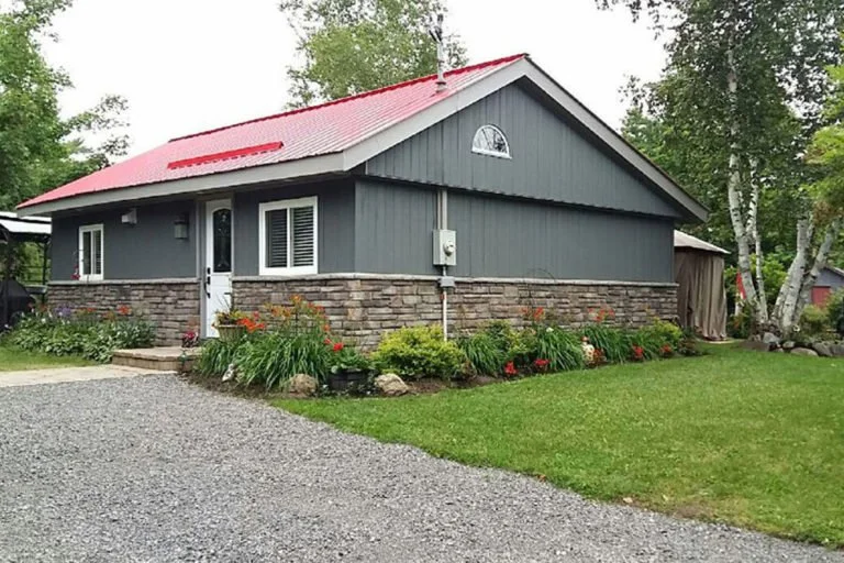 A gray house with a red metal roof, brick siding at the bottom, white window frames, surrounded by a green lawn and garden beds with colorful flowers.