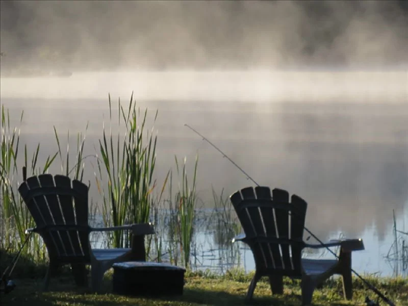 Two Adirondack chairs facing a misty lake, with fishing poles propped against the chairs and tall grass near the water.