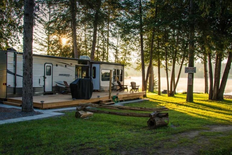 A camper trailer on a wooden deck near a lakeside with trees and sunlight at sunset.