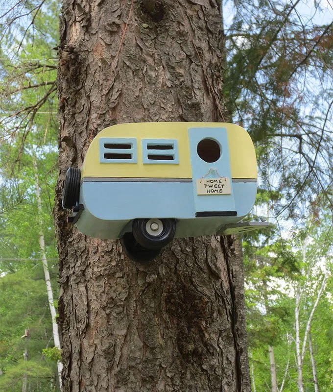 A camera designed to look like a vintage camper van is mounted on the trunk of a tree. The camera has a pastel yellow and blue color scheme with a round lens and two small vents. A small sign says 'Home Tweet Home'.