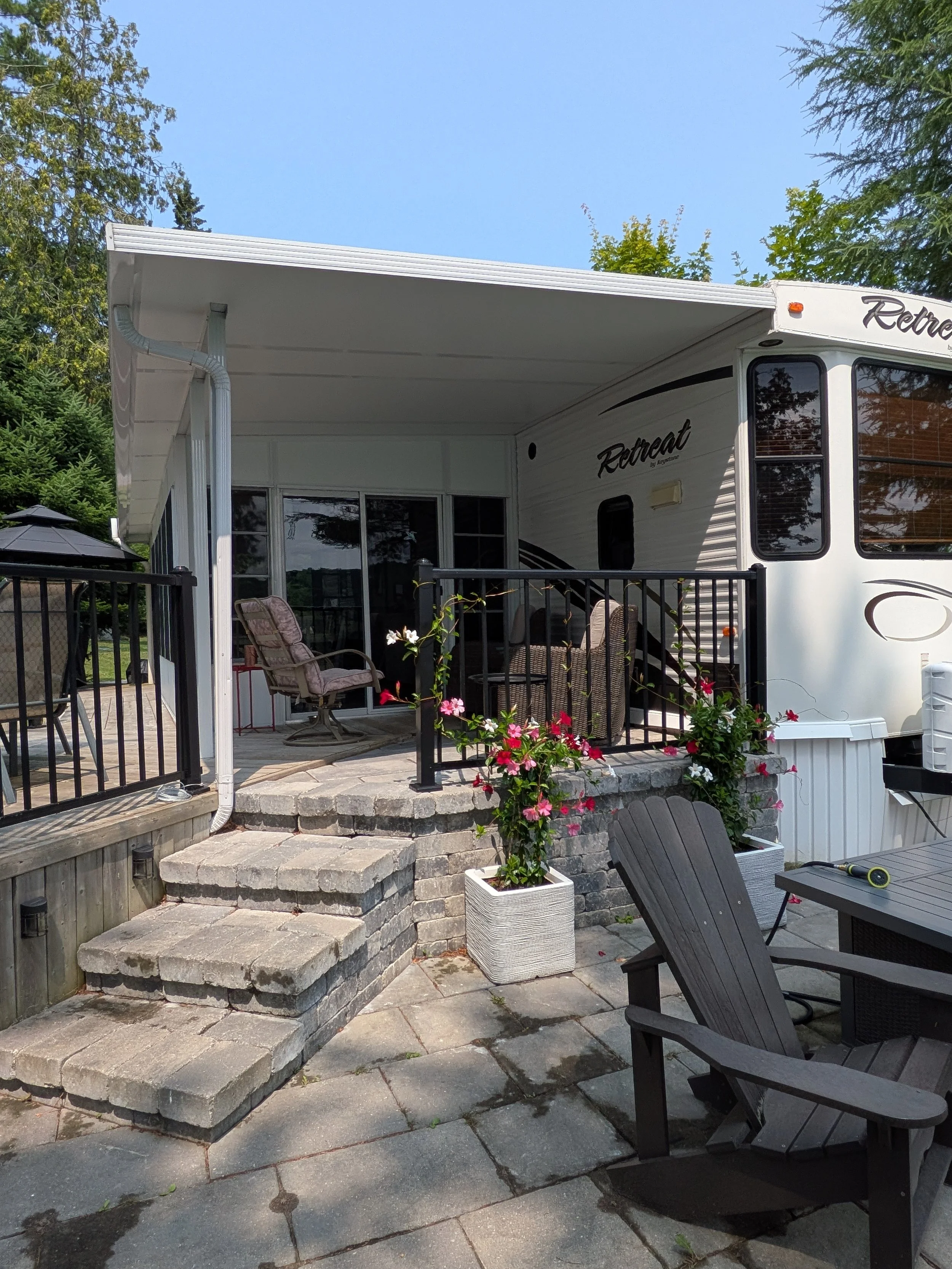 A backyard patio with a black metal railing, a set of stone steps leading to the patio, outdoor furniture including chairs and a table, potted plants, and a large RV trailer with a screened porch
