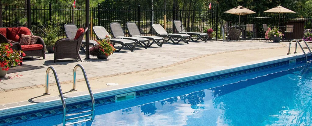 Empty swimming pool with lounge chairs and patio furniture on a deck surrounded by flowers and trees.