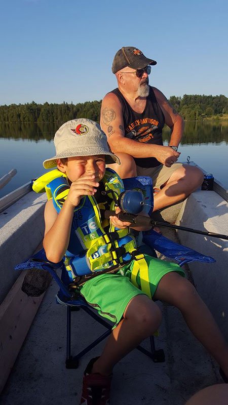 A young boy and an older man sitting in a boat on a lake, fishing. The boy is smiling while holding a fishing rod, wearing a hat and a life jacket. The man is wearing sunglasses, a sleeveless Harley-Davidson shirt, and a cap.