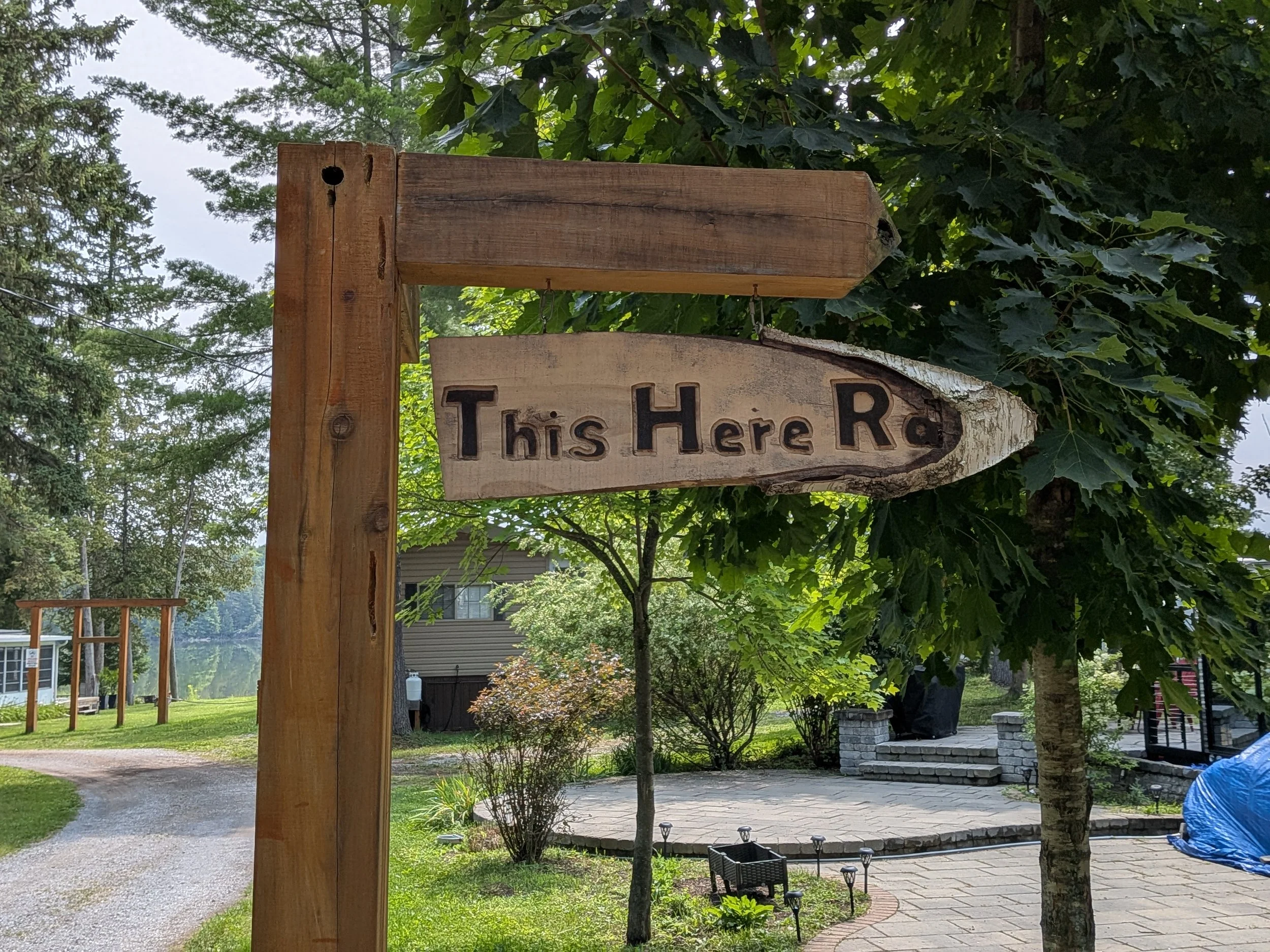 A wooden signpost in a backyard garden with the words 'This Here Ra' carved into it, surrounded by green trees and landscaping.