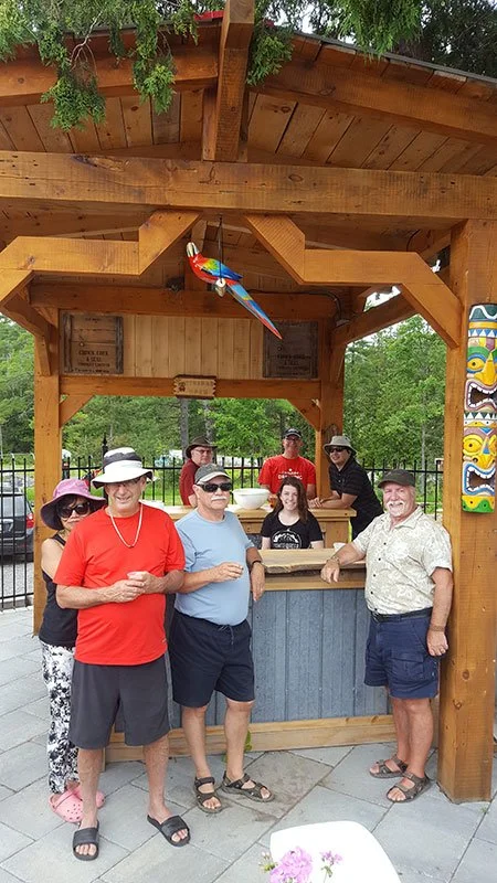 Group of seven people, five men and two women, is standing outside under a wooden pavilion with a colorful parrot hanging from the ceiling. They are dressed casually, some wearing sunglasses and hats, smiling at the camera. The background shows green