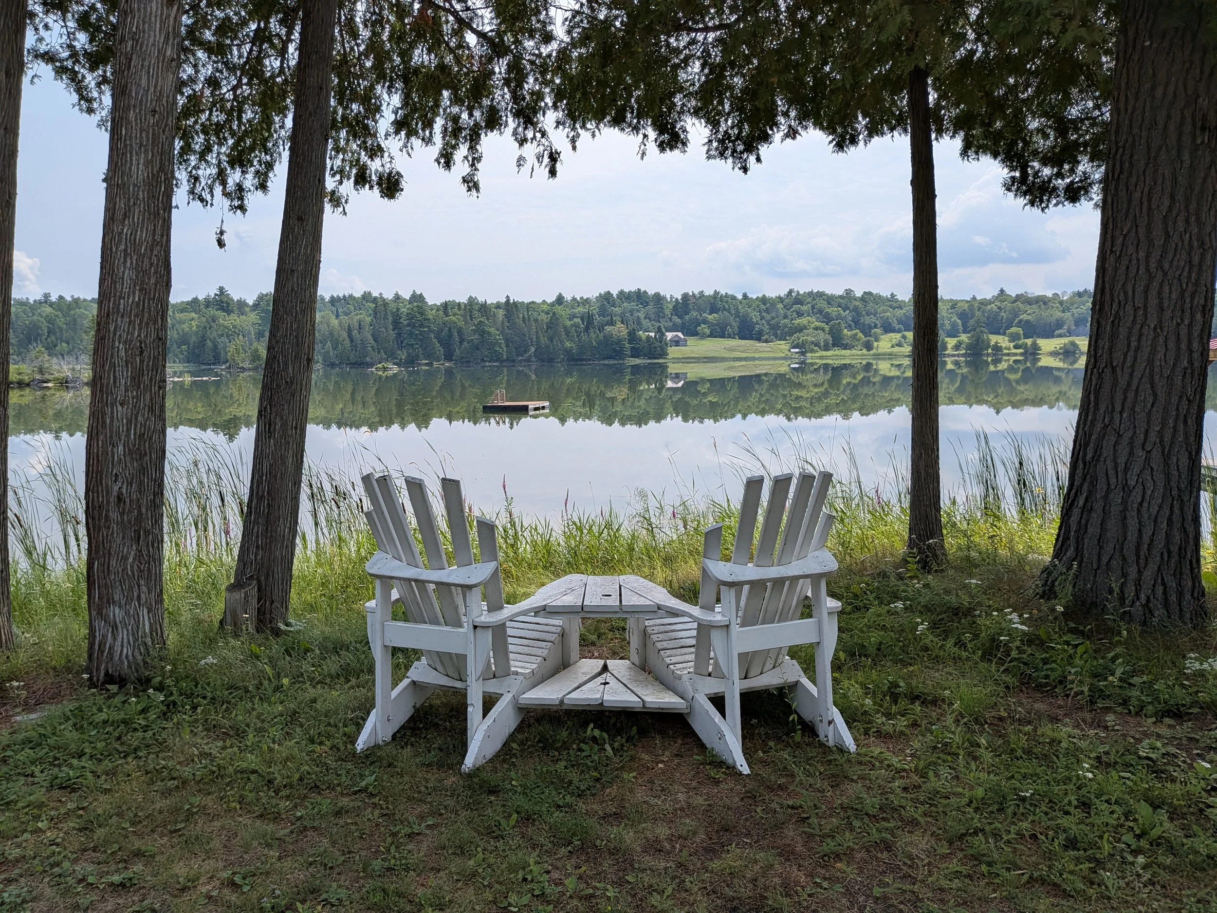White Adirondack chairs arranged in a circle near a lake, with trees and a dock reflected in the water in the background.