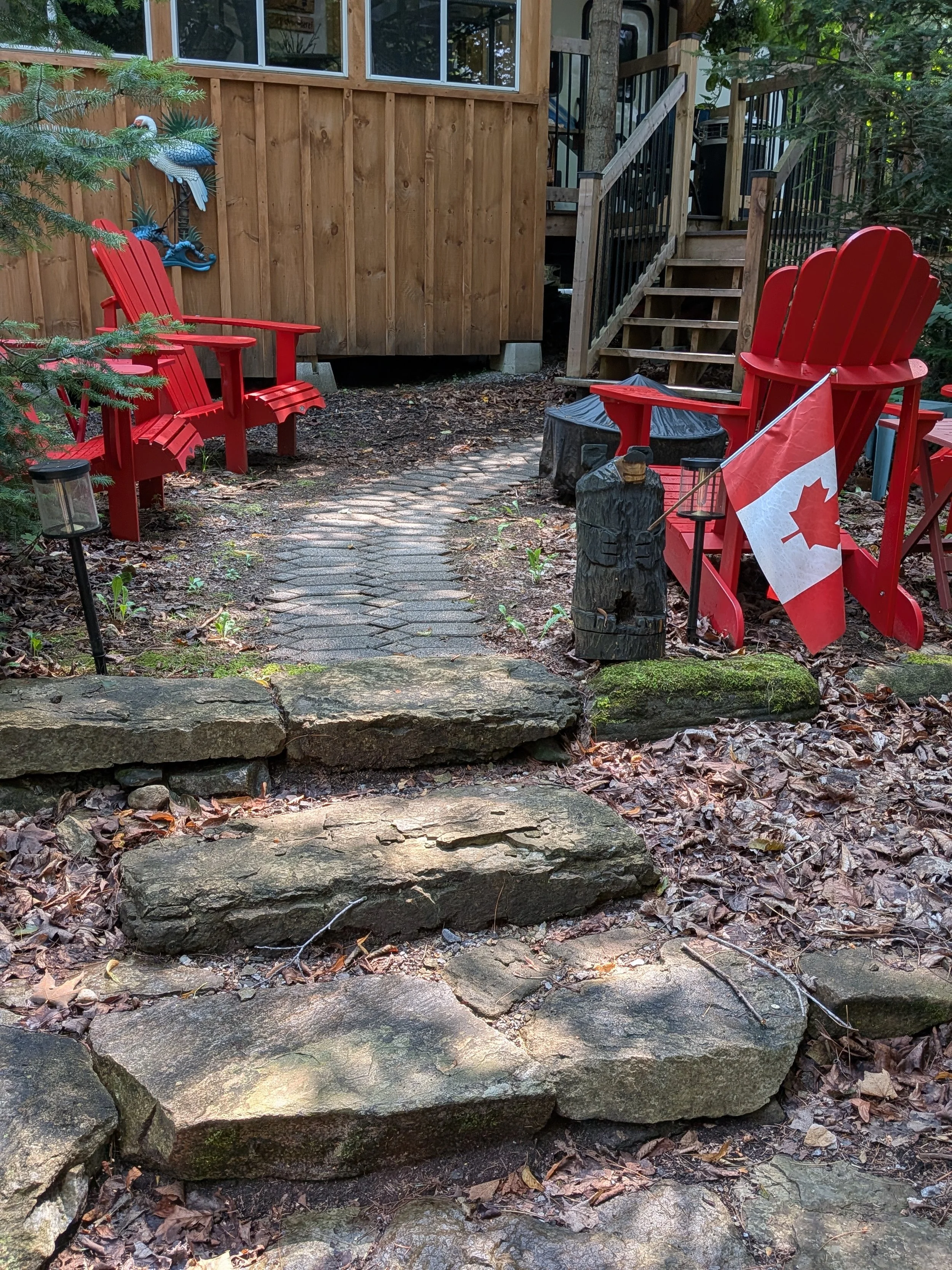 A backyard patio with a stone pathway, two red Adirondack chairs, a Canadian flag, a small fire pit, and decorative bird and dragonfly wall art near a wooden staircase leading to a house with a wooden exterior.