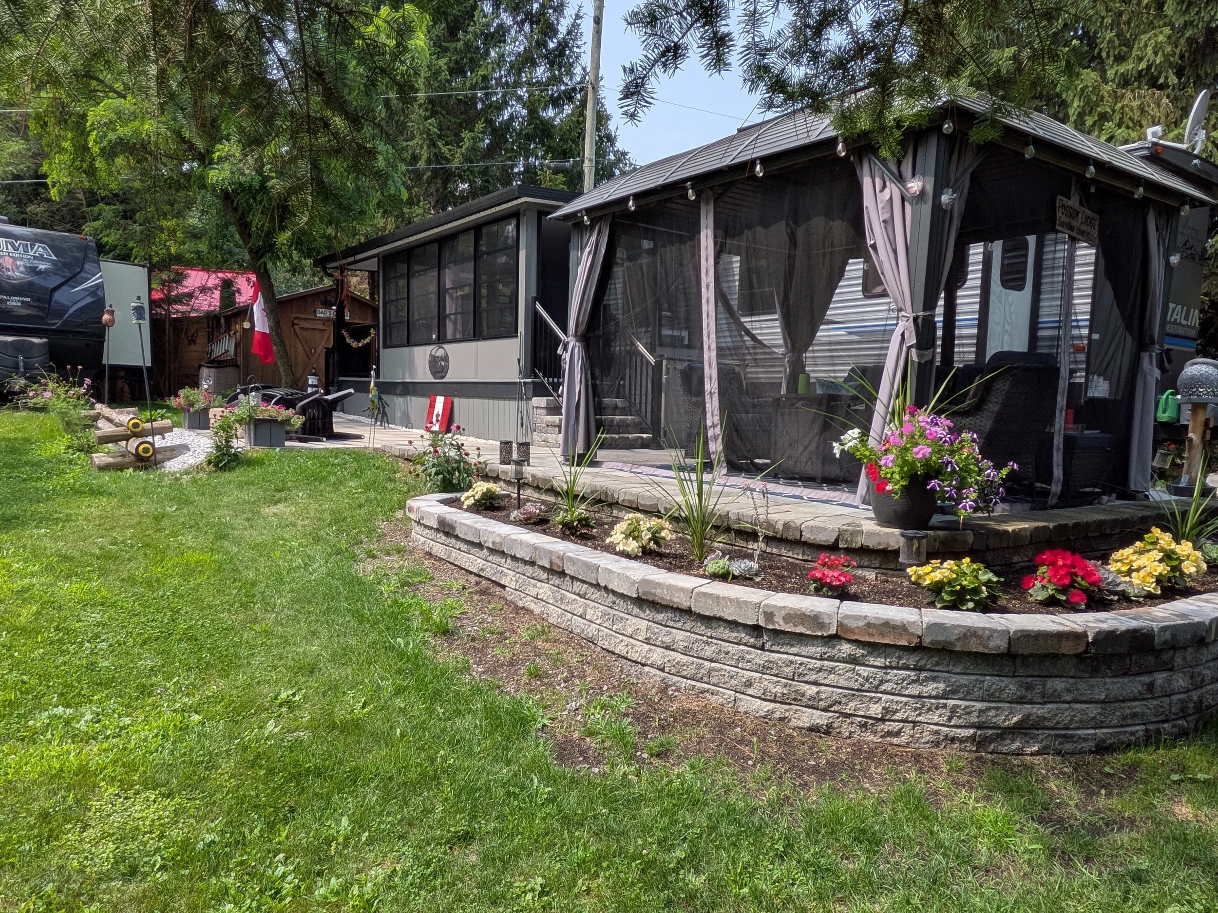 A backyard with a lawn, flowers, and a raised flower bed, featuring a large screened porch with curtains, plants, and outdoor furniture, along with trees and a power line overhead.