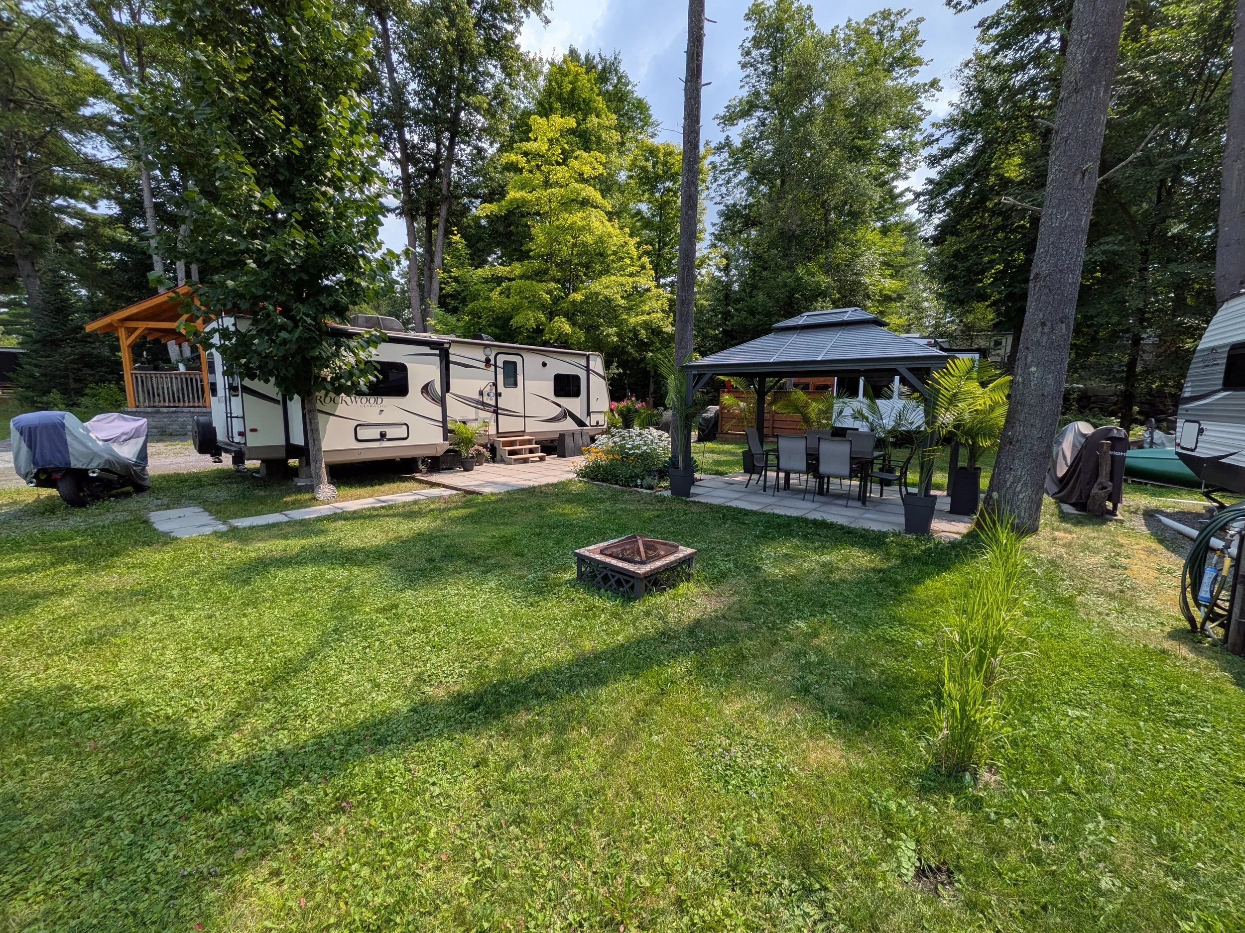 A backyard with green grass, trees, and outdoor furniture including a table with chairs under a gazebo, and a recreational vehicle. There is also a covered structure with a wooden roof and various outdoor items such as a grill and plants.