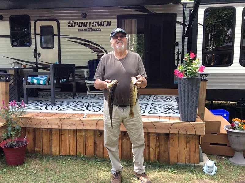 A man standing on a wooden deck holding two fish, one in each hand, outside a camper trailer.