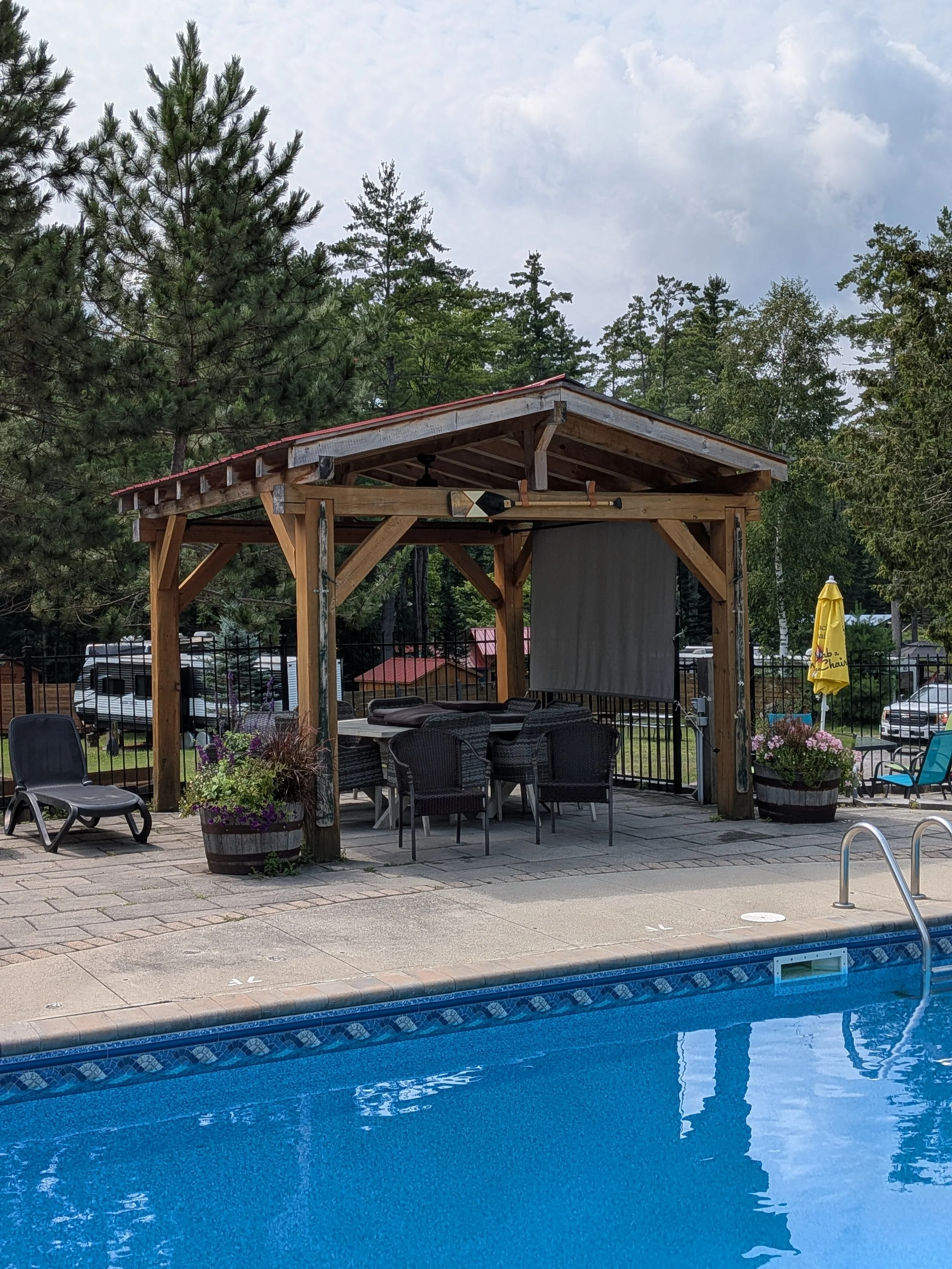 View of a backyard pool with blue water, surrounded by a concrete deck. There is a wooden pergola with outdoor furniture underneath, including a table and chairs. A yellow umbrella with the logo 'Chili's' is visible, along with potted flowers and out
