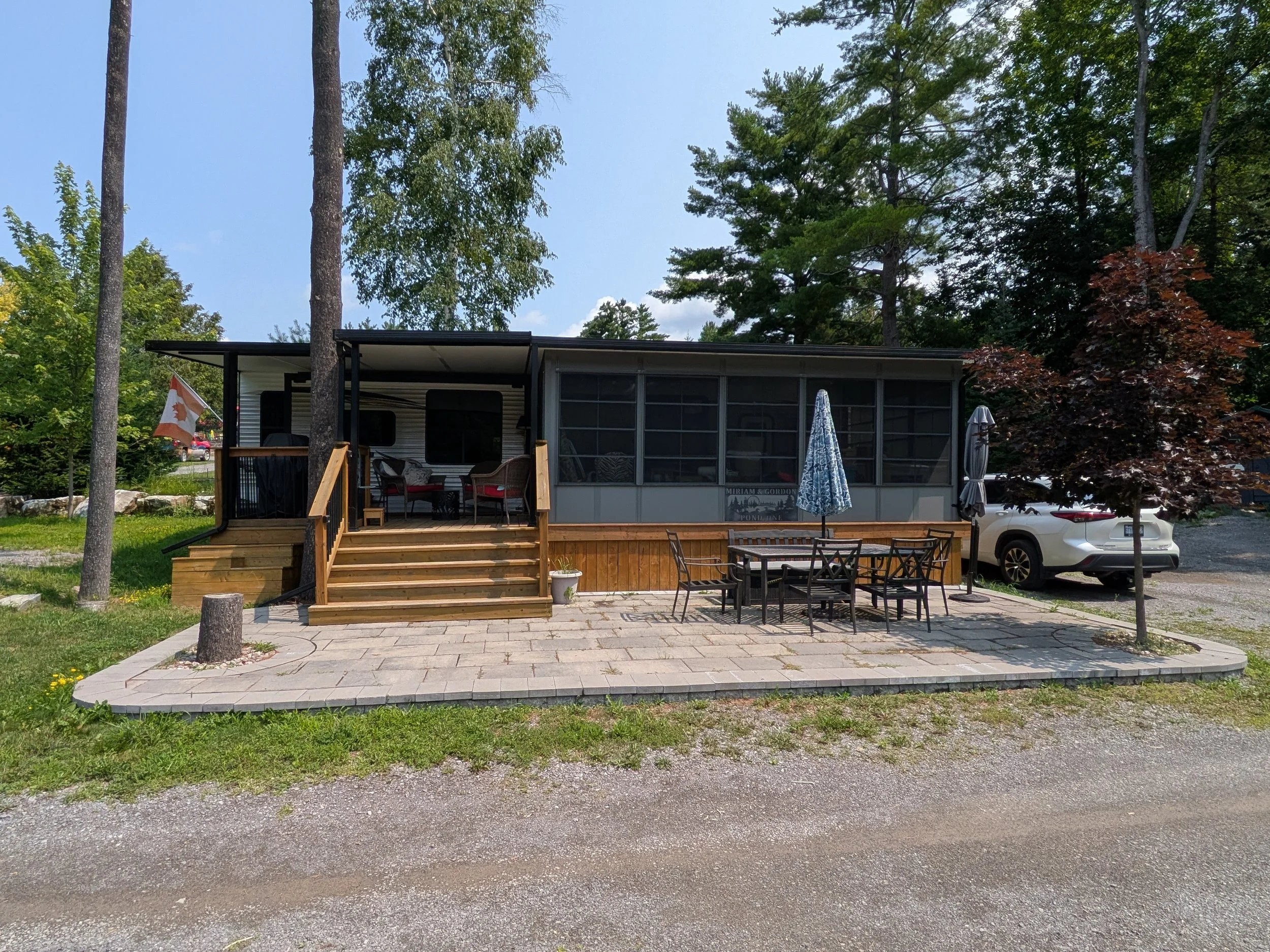 A mobile home with a screened porch, outdoor furniture including a table and chairs, umbrellas, and an Ontario flag, situated on a paved patio with trees and a car in the background.