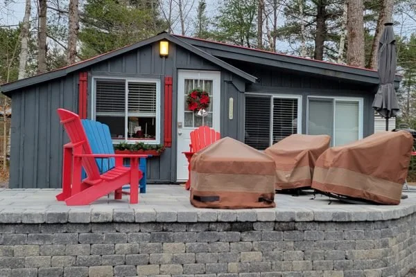 A gray house with red accents and a wreath hanging on the window, surrounded by outdoor furniture including red and blue Adirondack chairs and covered patio chairs, on a stone patio in a wooded area.