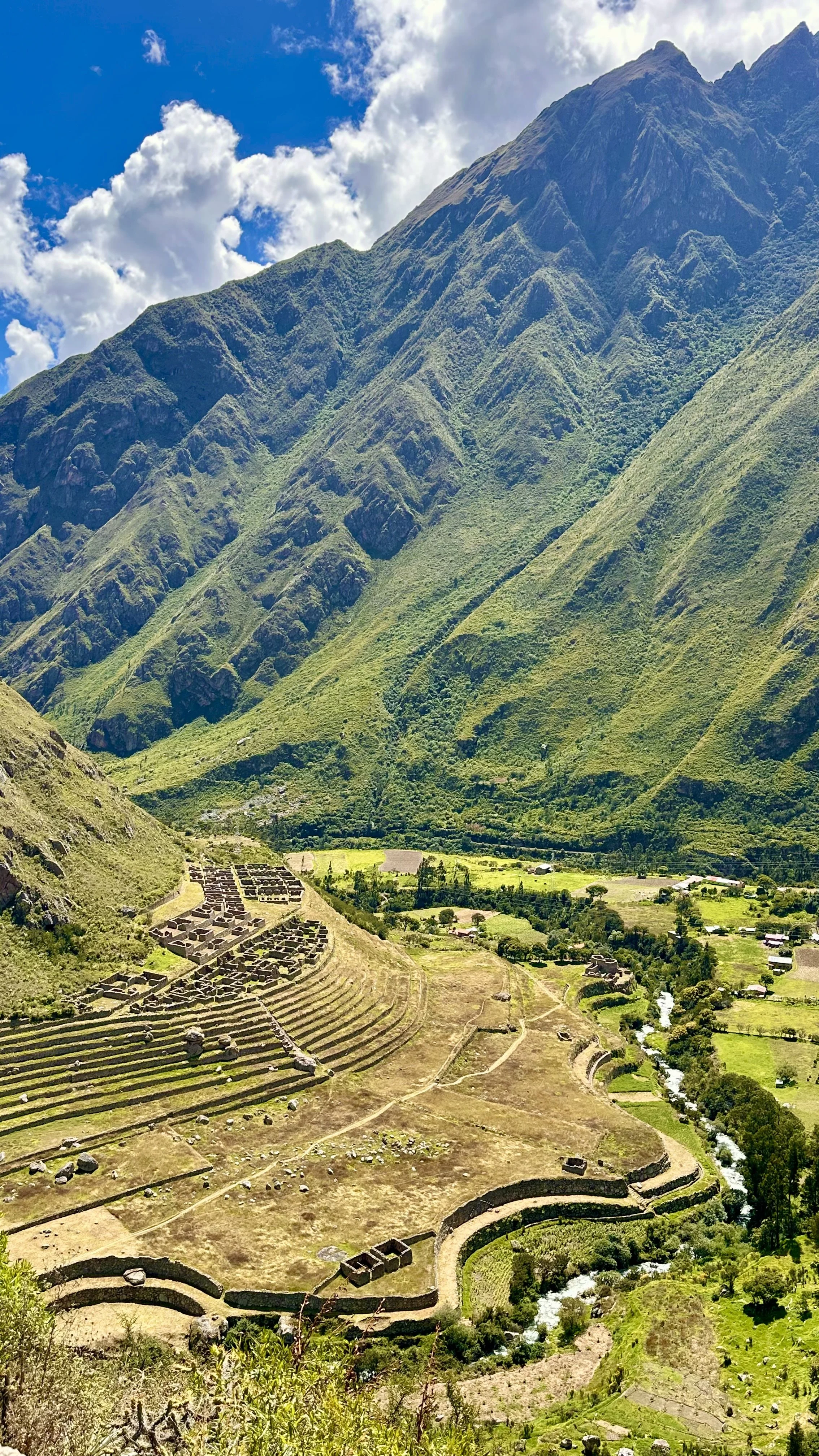 Ancient ruins in a lush green valley surrounded by tall, rugged mountains with a partly cloudy sky overhead.