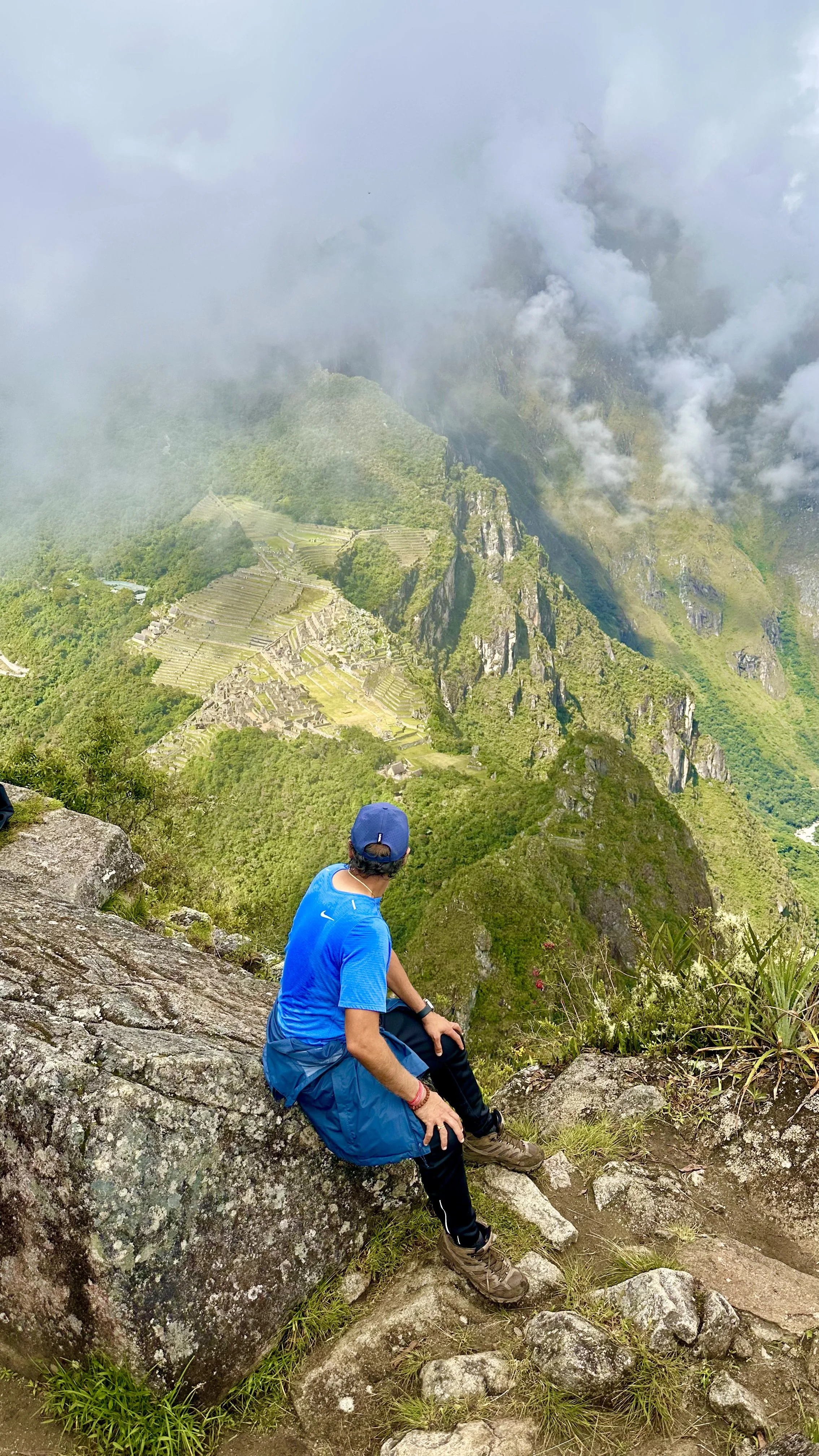 A person sitting on a rocky ledge overlooking Machu Picchu, surrounded by clouds and lush green mountainous terrain.