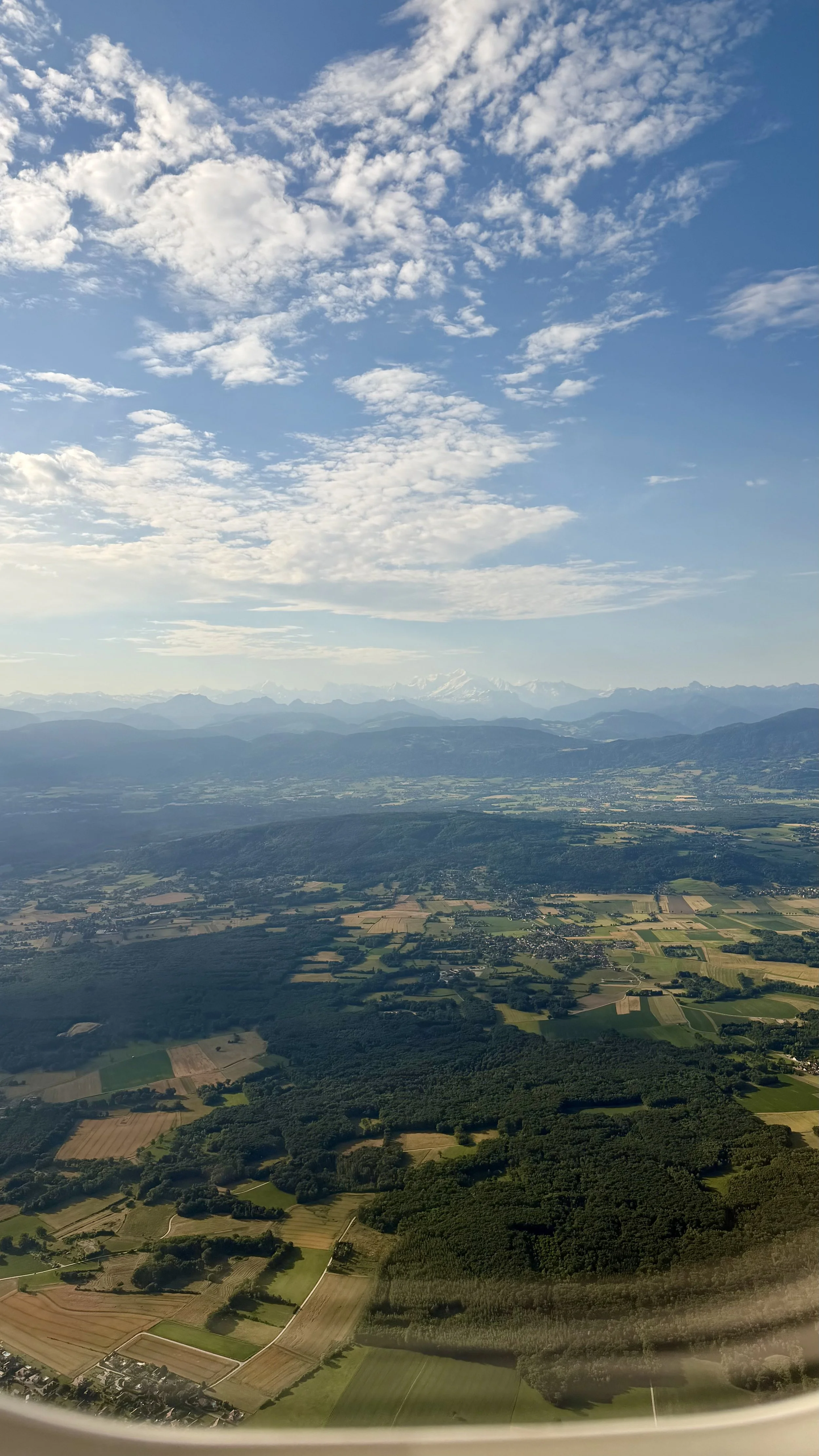 Aerial view of a landscape with fields, forests, mountains, and a partly cloudy sky.