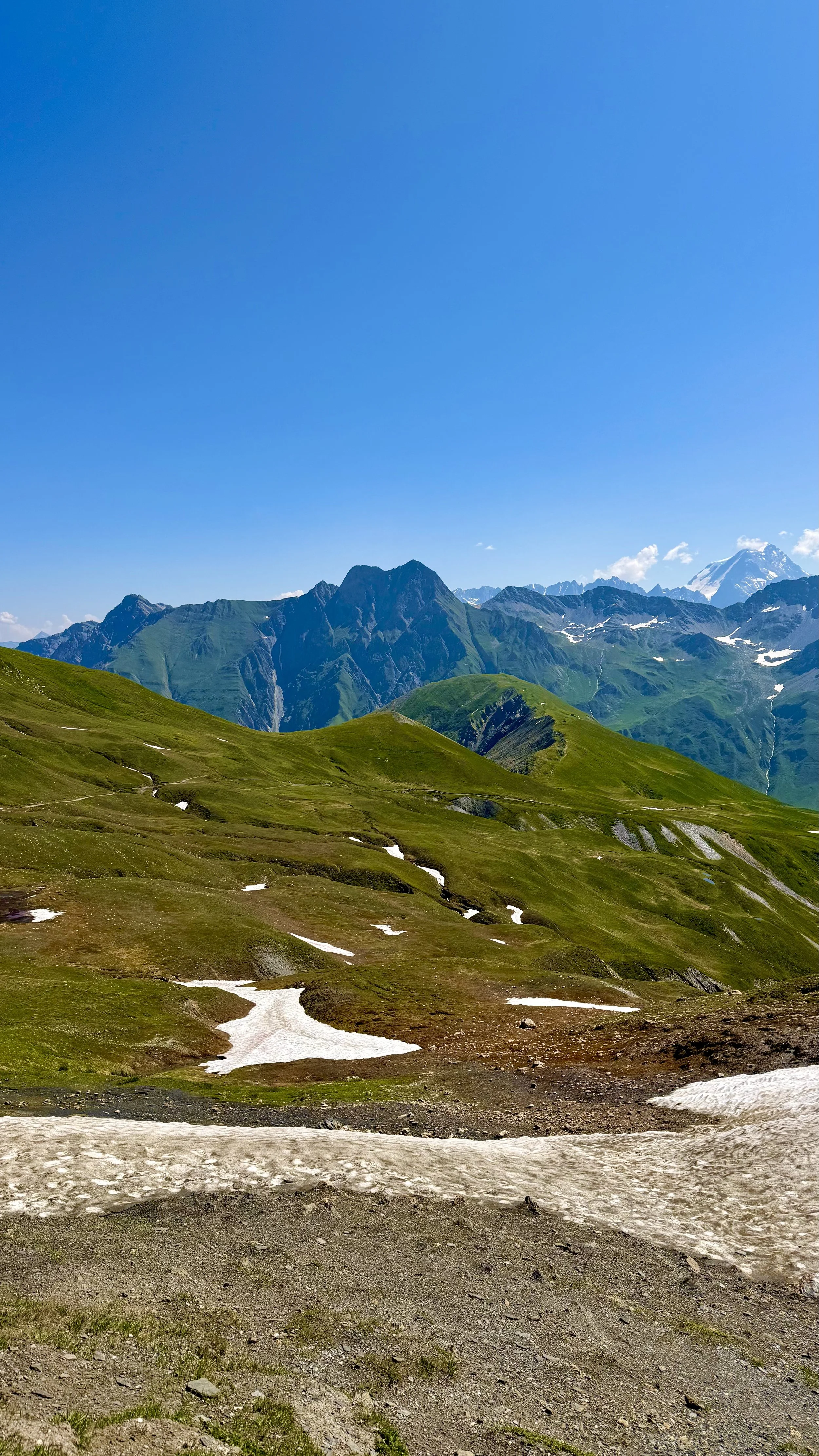 Scenic mountain landscape with green rolling hills, snow patches, tall mountains in the background, and a clear blue sky.