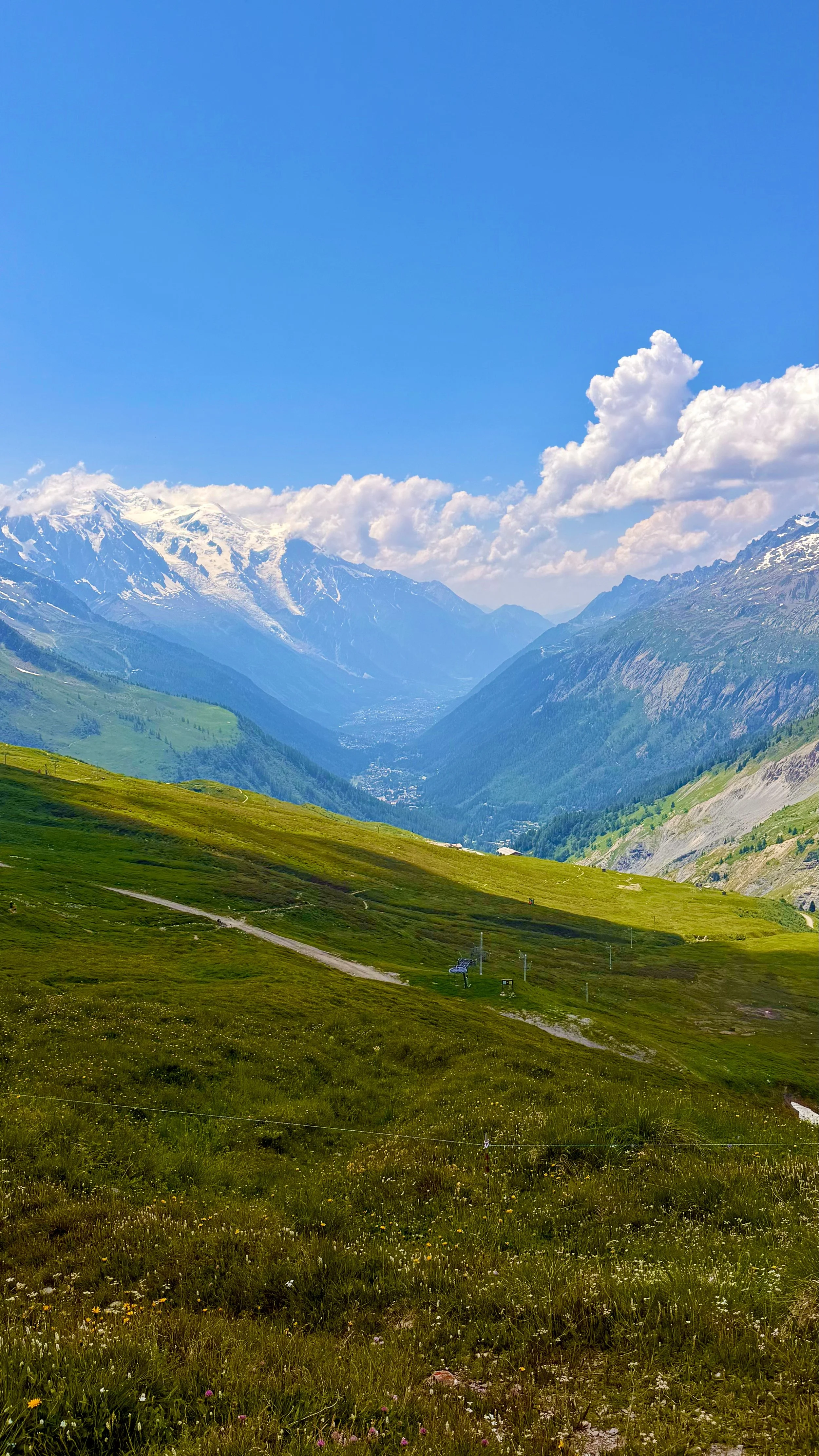 Scenic view of a green valley with mountainous terrain and snow-capped peaks in the background, under a partly cloudy blue sky.