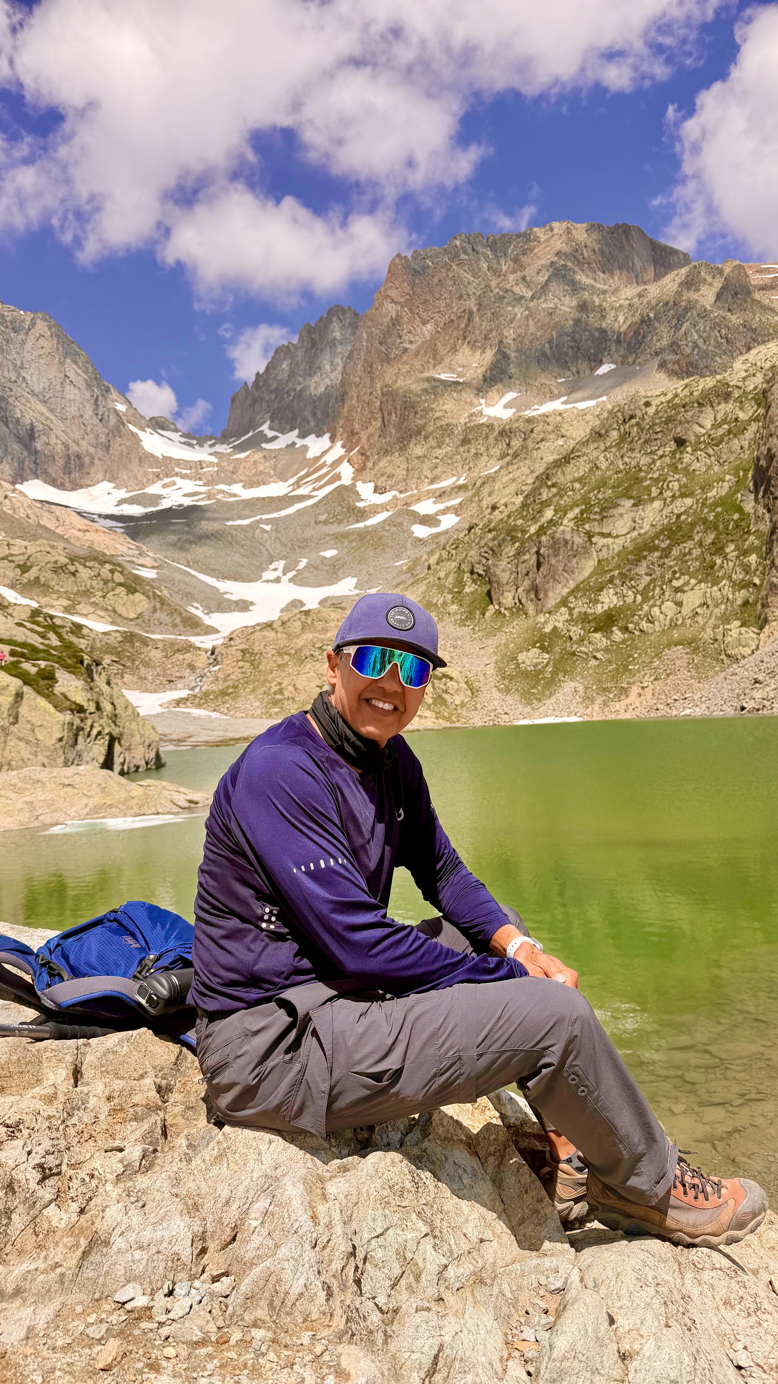 A man wearing a purple jacket, gray pants, hiking boots, and reflective sunglasses, sitting on rocks near a green alpine lake with snow-capped mountains and a partly cloudy sky in the background.