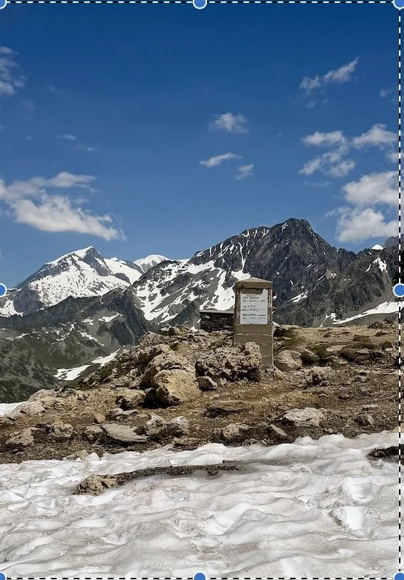 Snow on the ground with rocks and a signpost in the foreground. Mountain peaks with snow and dark rocky terrain in the background, under a blue sky with some clouds.
