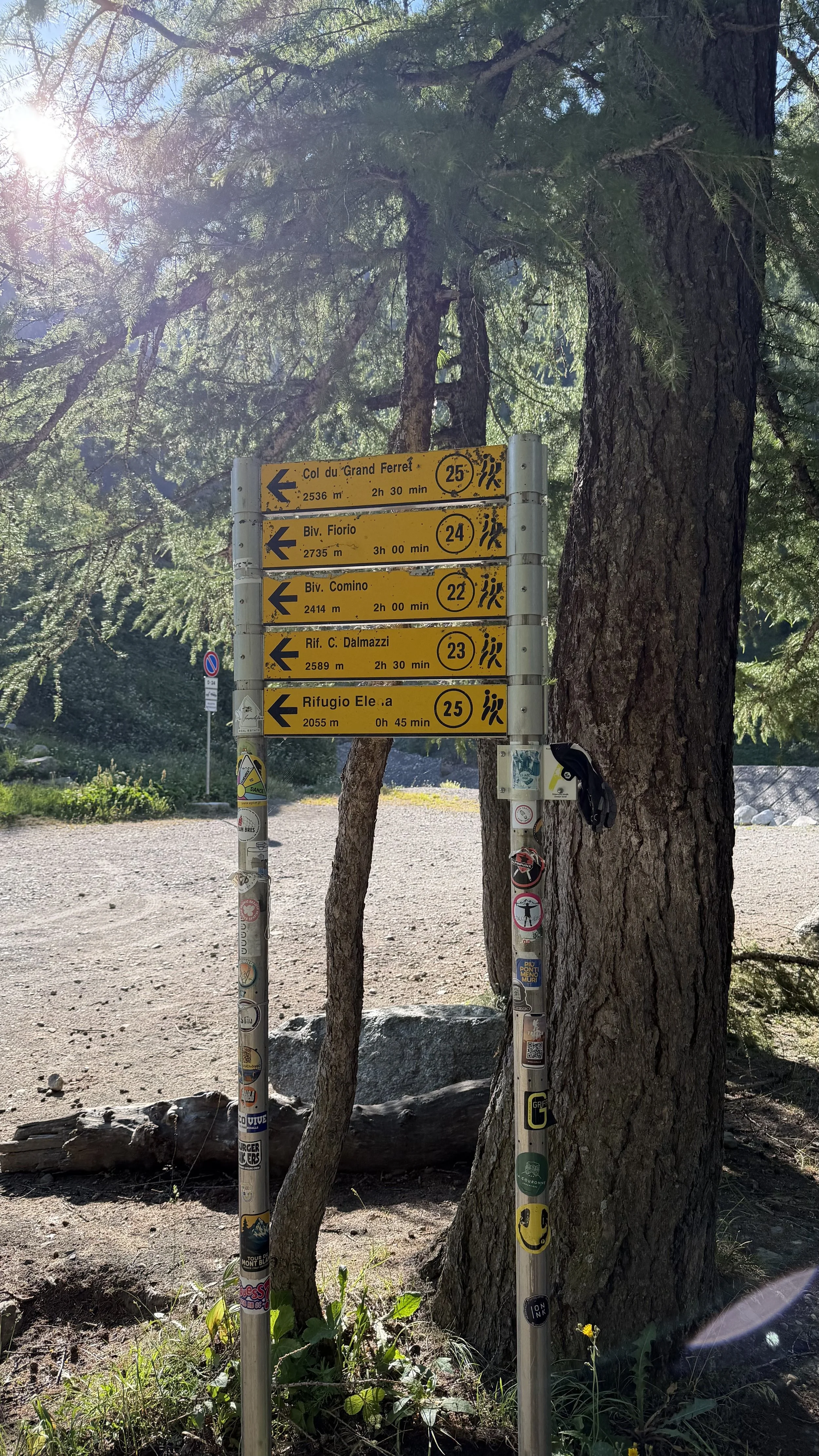 Trail signpost with directions and distances for various mountain locations, placed beside trees in a forested area on a sunny day.
