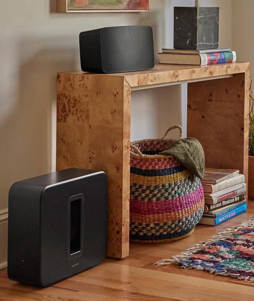 A wooden console table with a black speaker on top, a dark marble lamp, and a stack of books. A woven basket with a blanket inside is underneath the table, next to a pile of books and a colorful rug on a wooden floor.