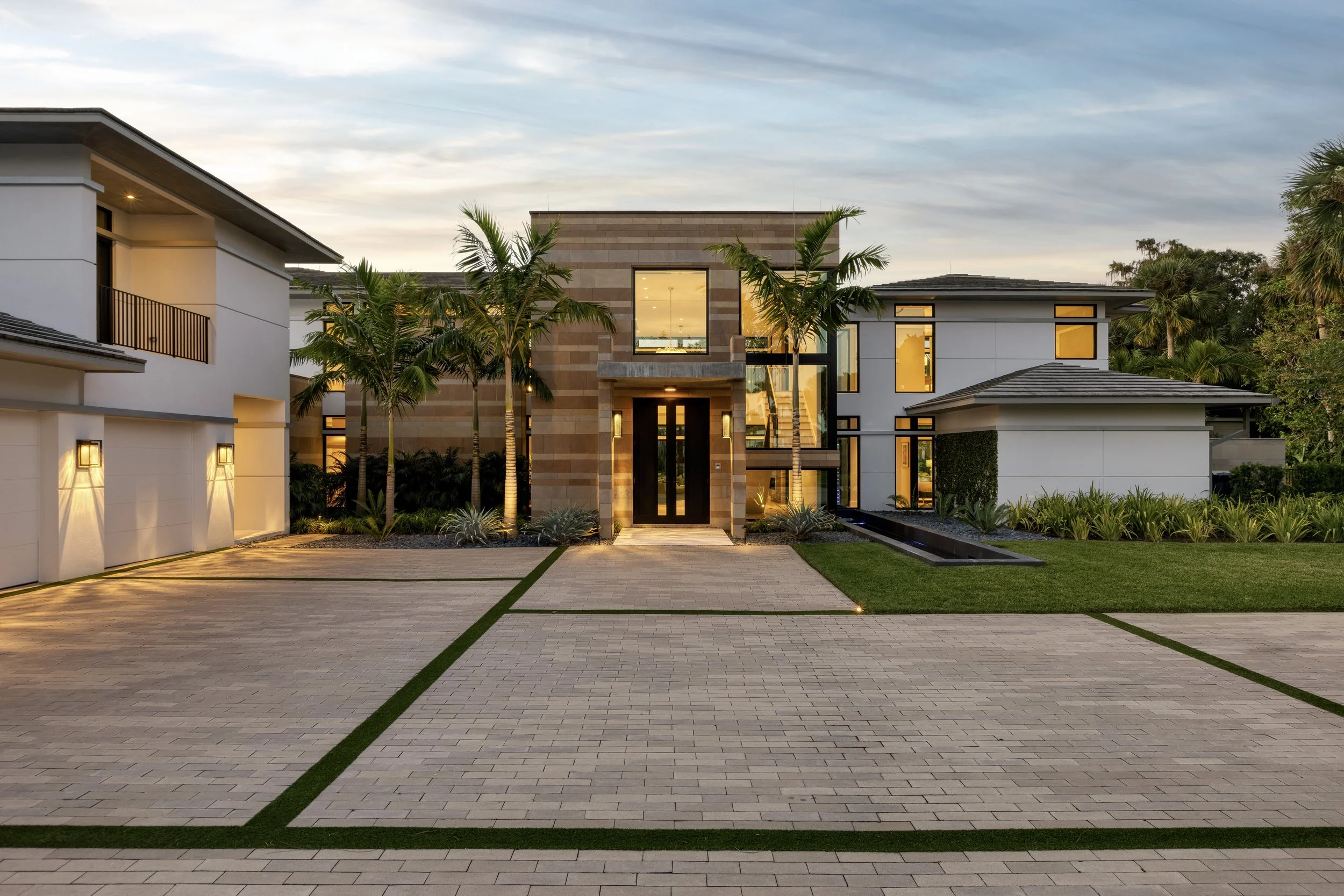 Modern residential house with a driveway, palm trees, and landscaped yard at dusk, with lights on inside.