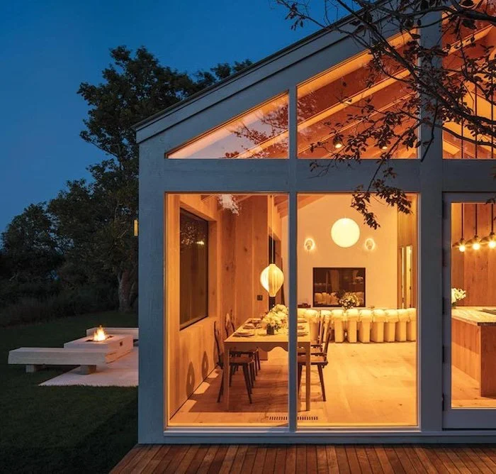 Interior view of a modern house with large glass windows, showing a dining area with a wooden table, chairs, and warm hanging lights, and a cozy living space with a fireplace and decorative lighting, during evening or dusk.