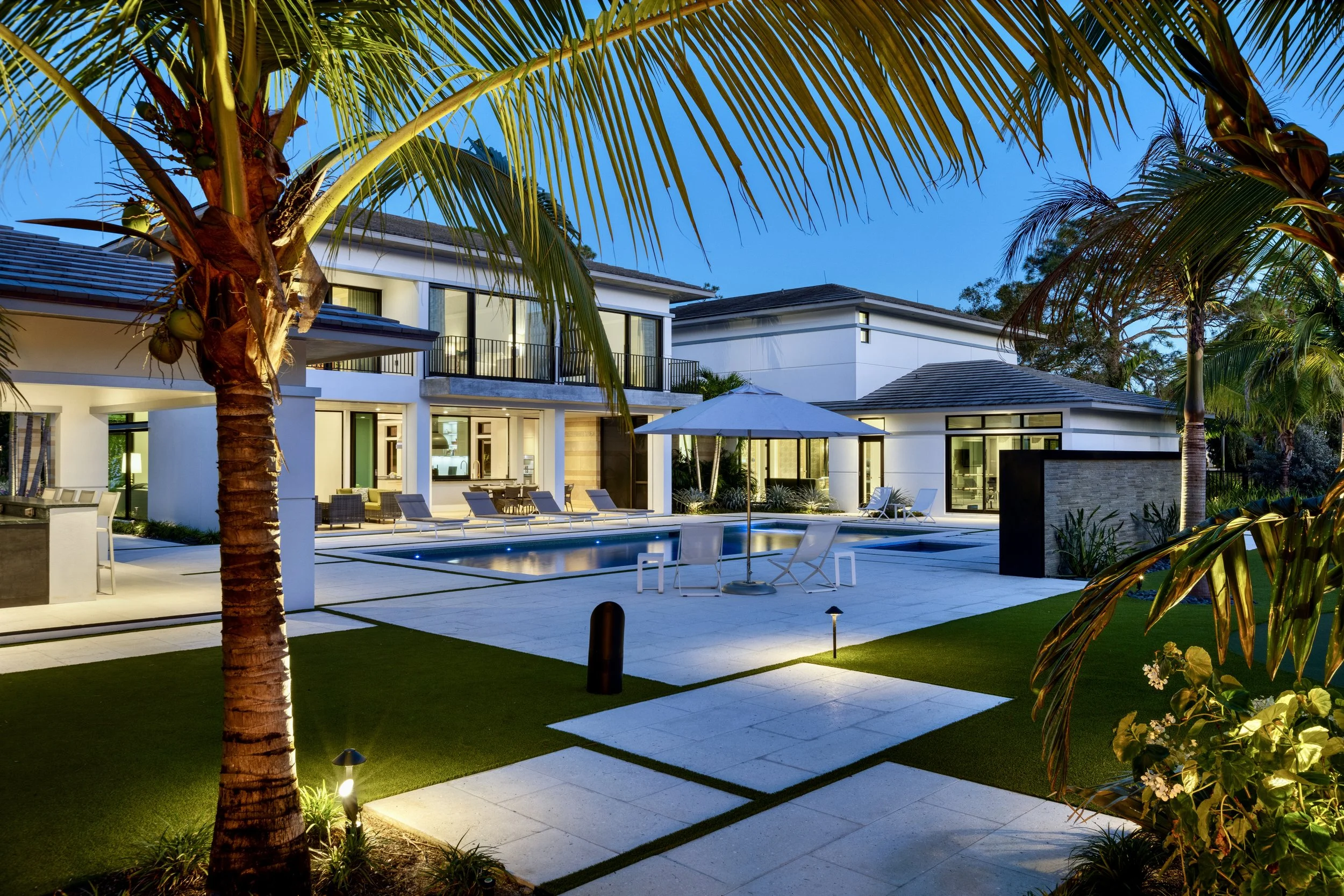 Modern house with backyard pool, lounge chairs, and umbrellas, surrounded by palm trees and illuminated outdoor lighting at dusk.