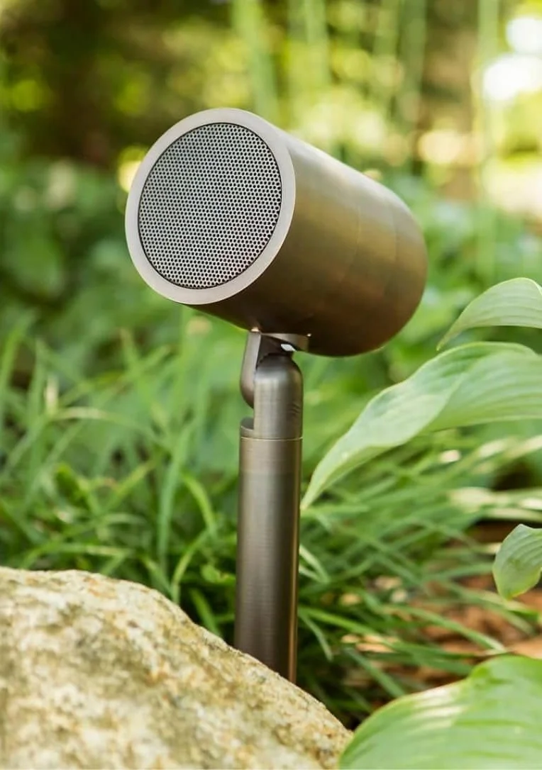 Outdoor speaker mounted on a metal post among green plants and rocks in a natural setting.