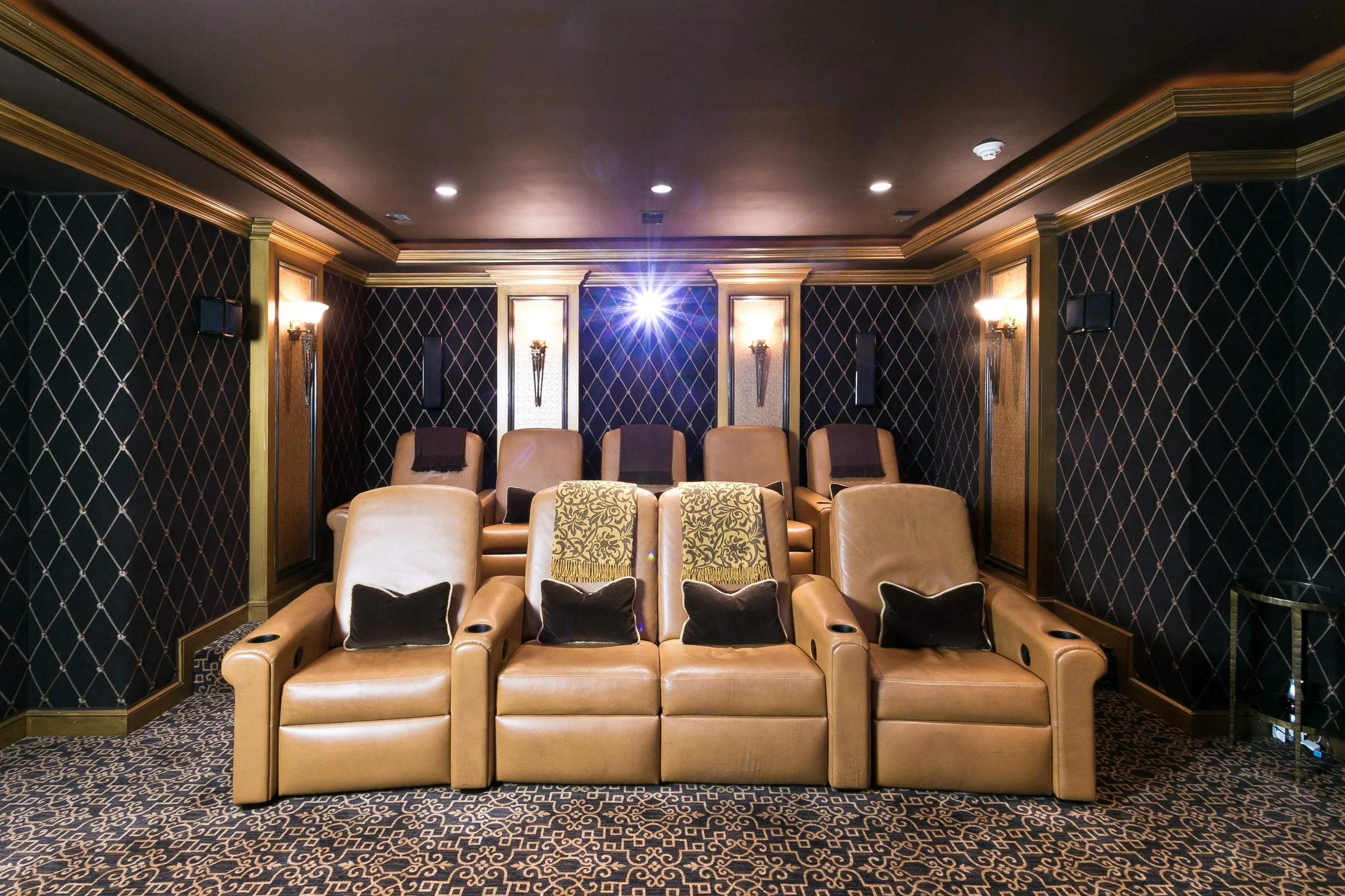 Luxury home theater with leather recliner seats, black and gold ornate wallpaper, decorative wall sconces, and a projector shining light on the ceiling.