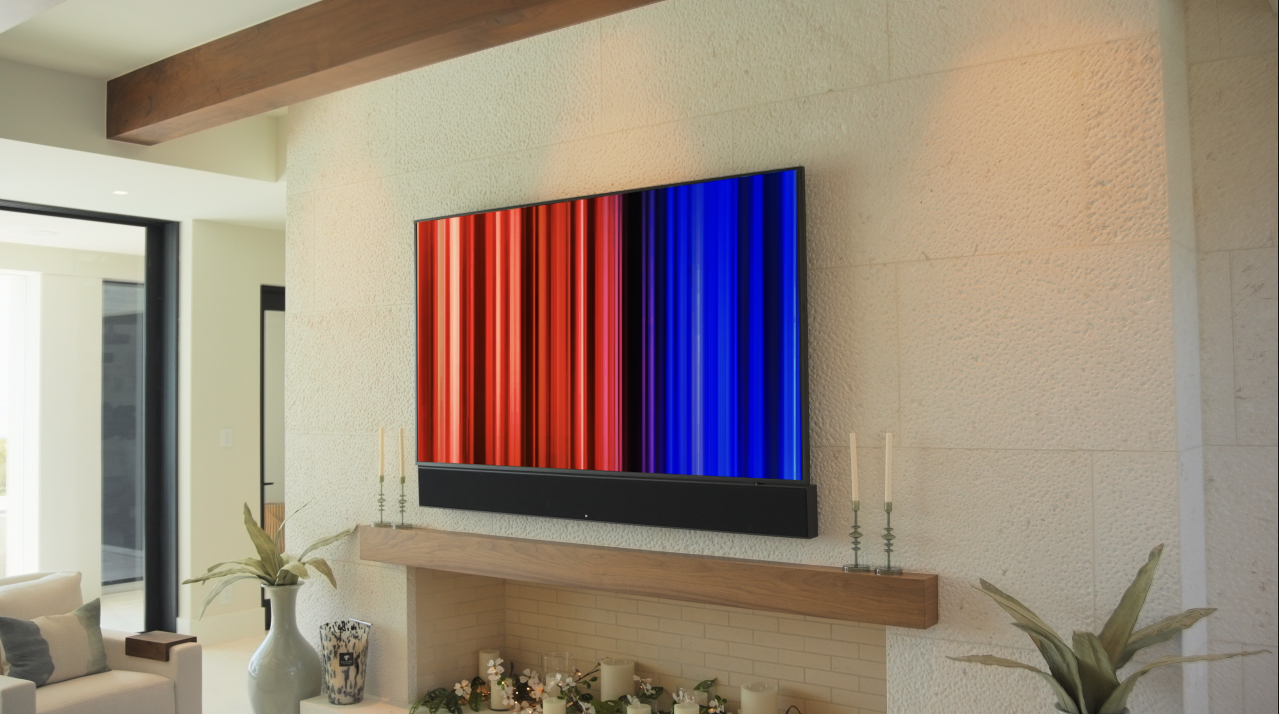 A modern living room with a mounted flat-screen television displaying a colorful vertical striped pattern. The room features a beige textured wall, a wooden mantel, a white vase with a plant, and decorative candles.