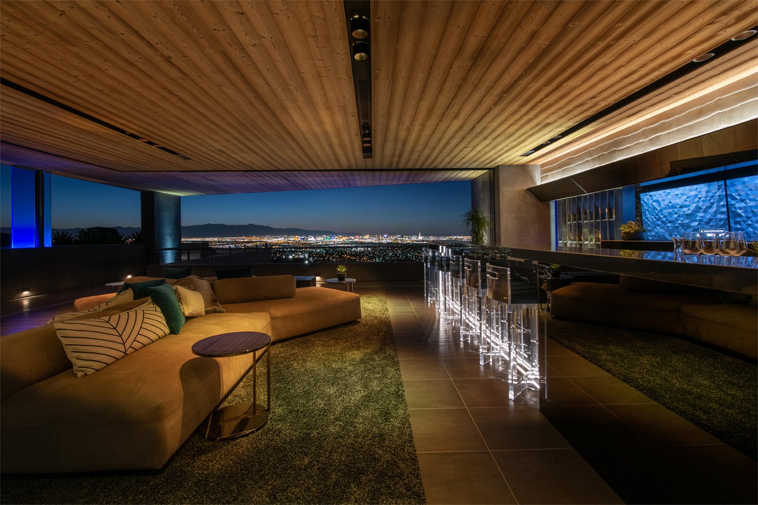 Luxurious living room with panoramic city view at night, featuring a beige sectional sofa with pillows, a small side table, a bar area with illuminated clear acrylic barstools, wood-paneled ceiling, and accent lighting.