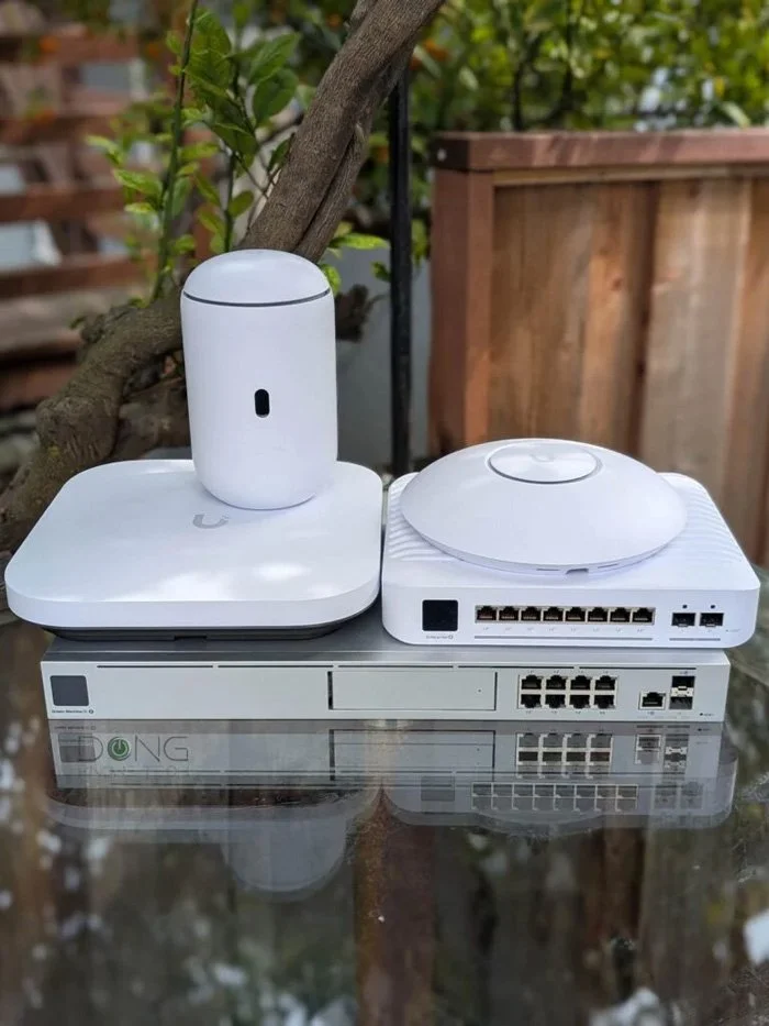 Stack of networking devices on a glass table outdoors, including a white router, a network switch, and a wireless access point.