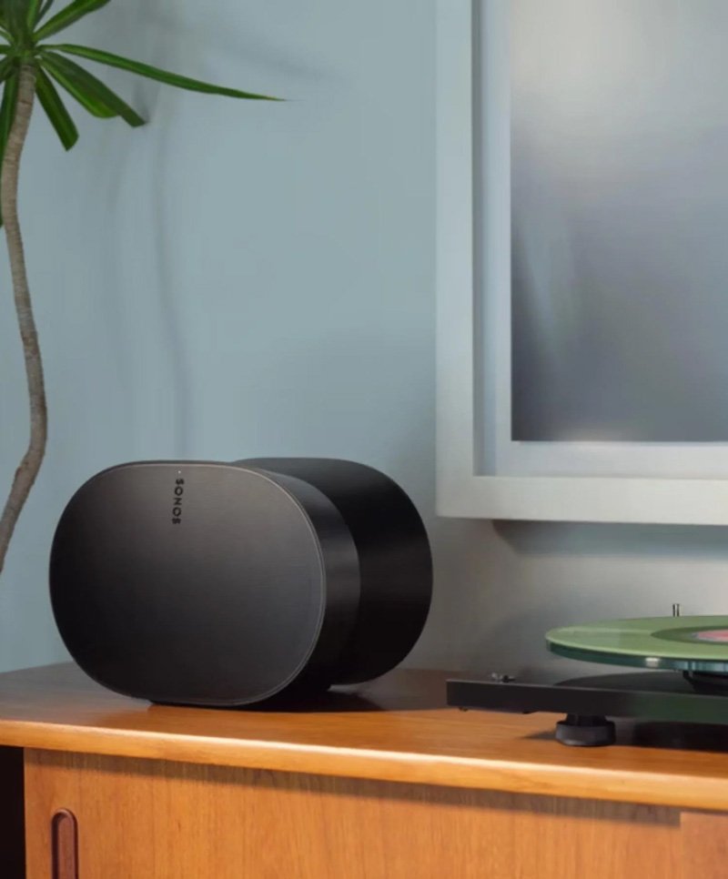 Black Sonos speaker on a wooden dresser next to a turntable with green records, with a plant and a computer monitor in the background.