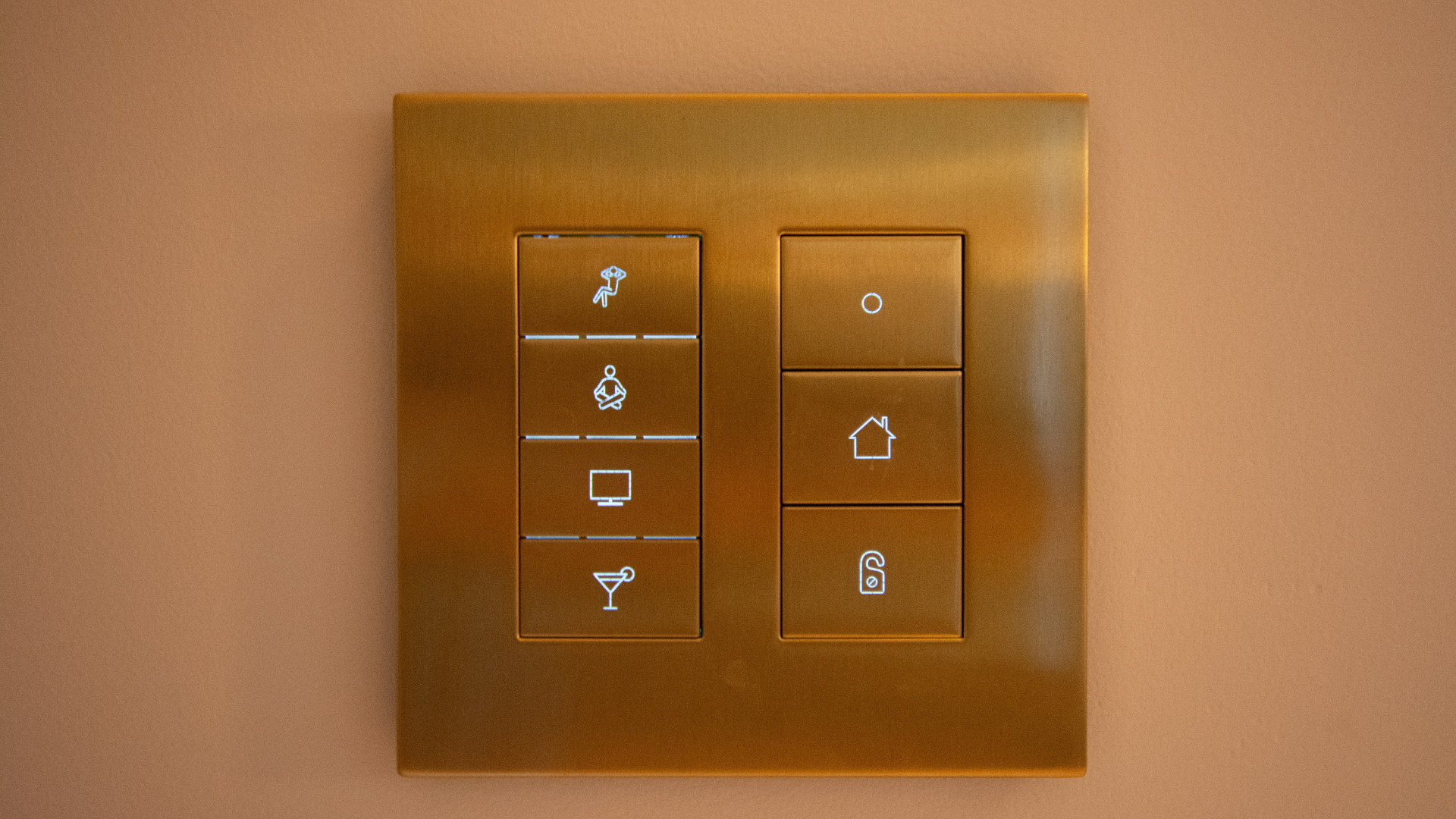 Gold-colored wall switch with illuminated icons for lighting control, including a lamp, sitting person, TV, drink, home, padlock, and doorbell.