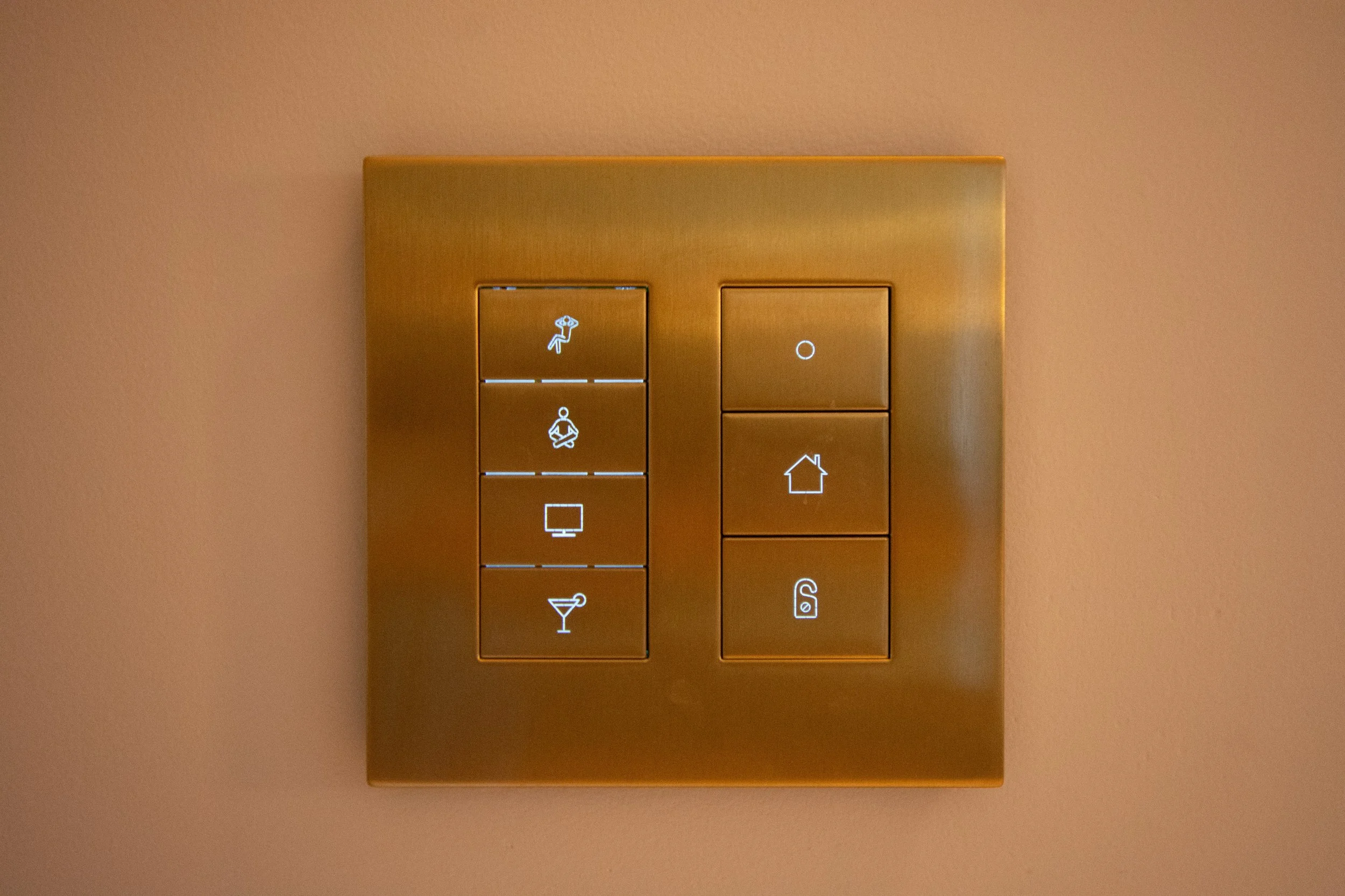 A close-up view of a gold-colored wall switch panel with illuminated icons, including a rain shower, a person meditating, a television, and a house.