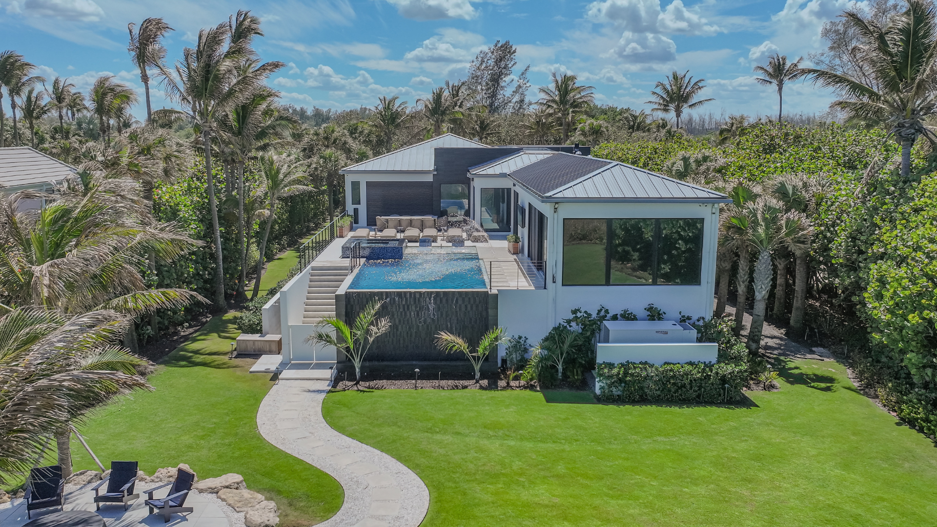 Modern house with a rooftop pool and outdoor seating area, surrounded by tall palm trees and lush green lawn under a partly cloudy sky.