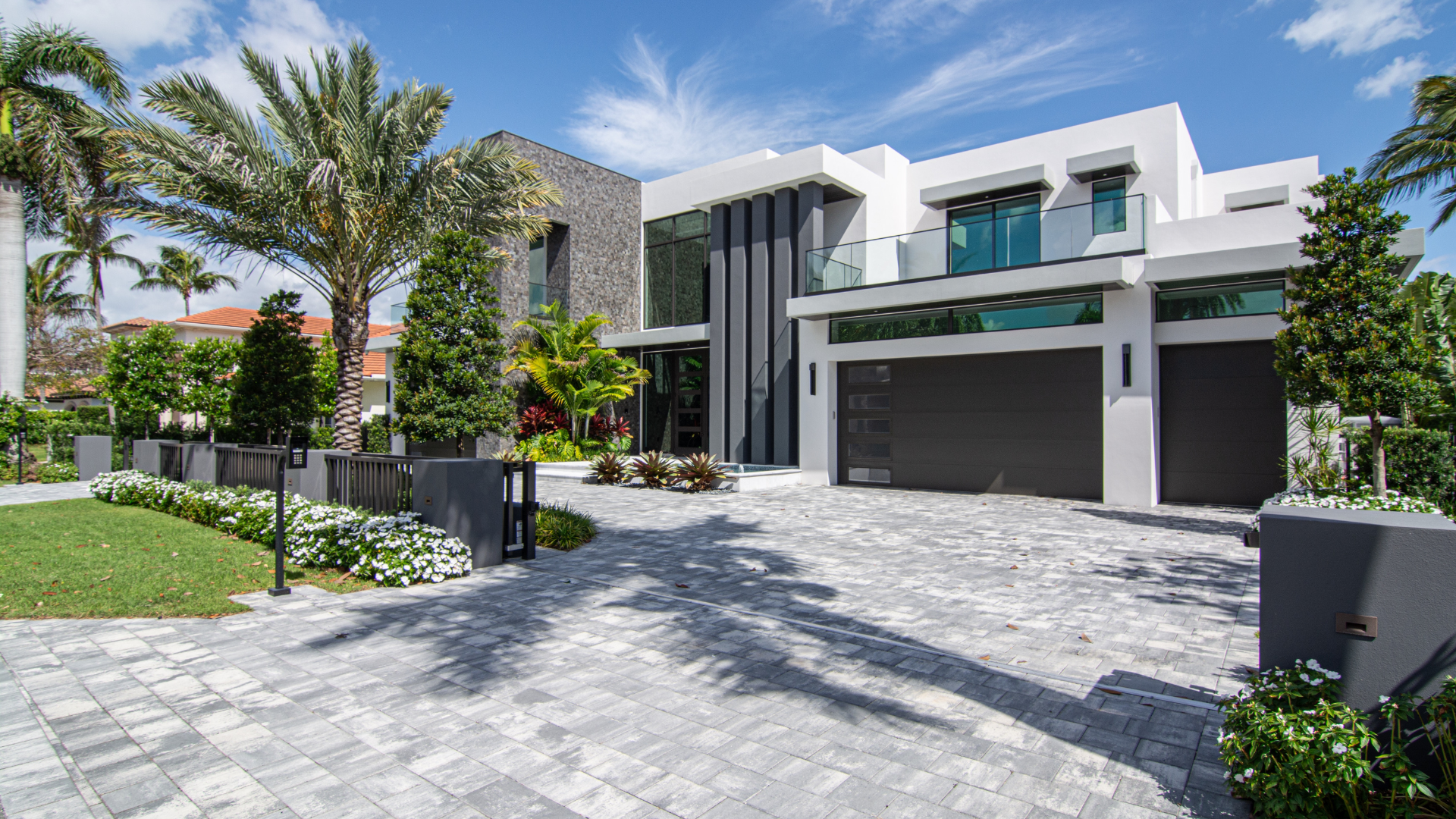 Modern Jupiter white house with black accents, large glass windows, and a garage, surrounded by lush greenery and palm trees under a blue sky with scattered clouds.