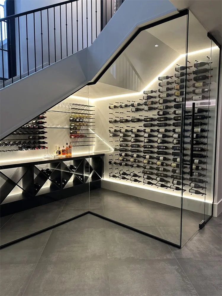Modern wine storage closet with glass walls, wine bottles on racks, and some liquor bottles on a counter, under a staircase with black railing and LED lighting.