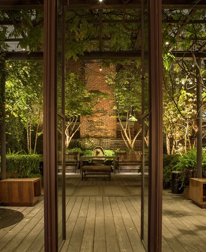 Nighttime view of a cozy outdoor patio with wooden flooring, lush green trees, potted plants, and seating areas, framed by glass doors.