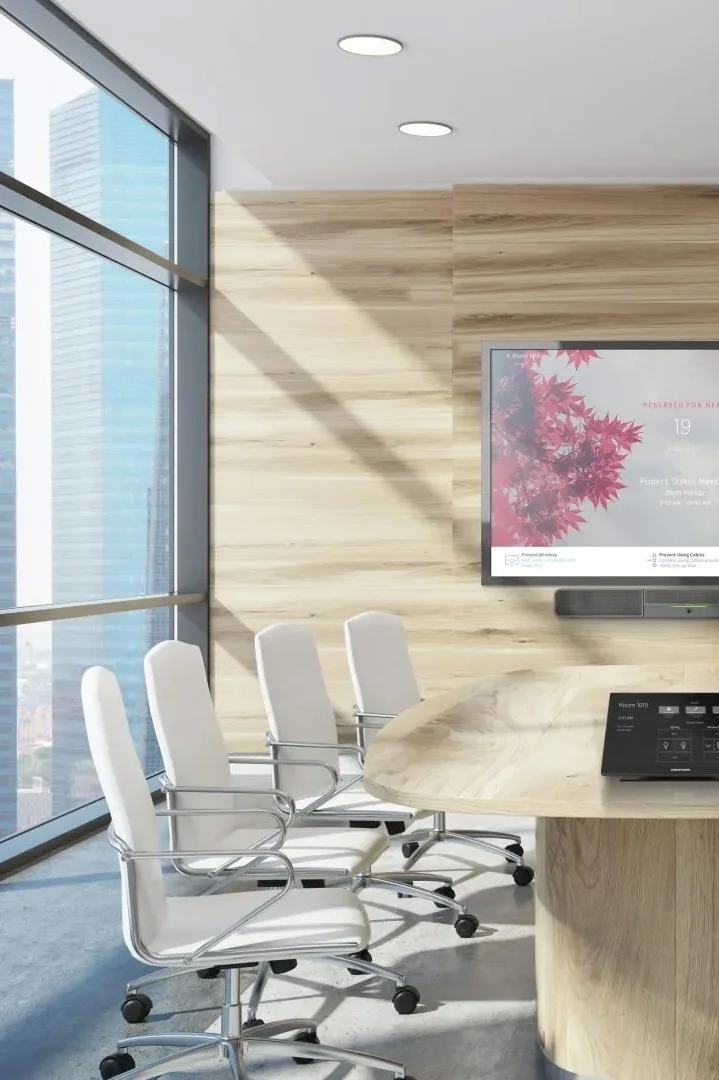 Modern conference room with white chairs and a large wooden table, large windows showing a city skyline, wall-mounted TV displaying a red leaf image, and ceiling lights.