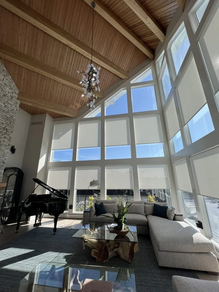 Bright living room with floor-to-ceiling windows, a wooden ceiling with exposed beams, a chandelier, a black grand piano, a light-colored sectional sofa with pillows, a glass coffee table with a decorative item, and a stone wall.