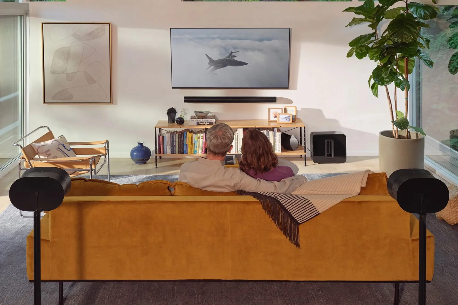 A couple sitting on a mustard-colored couch in a modern living room, watching TV. The room has large windows, a bookshelf with colorful books, a large potted plant, and minimalist artwork on the walls.