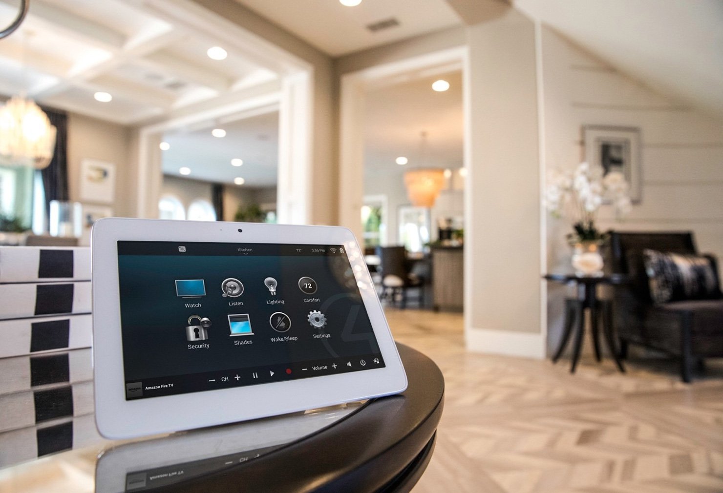 Smart home control tablet on a dark round table inside a bright living room with beige walls, hardwood floors, and white trim, showing various control options.